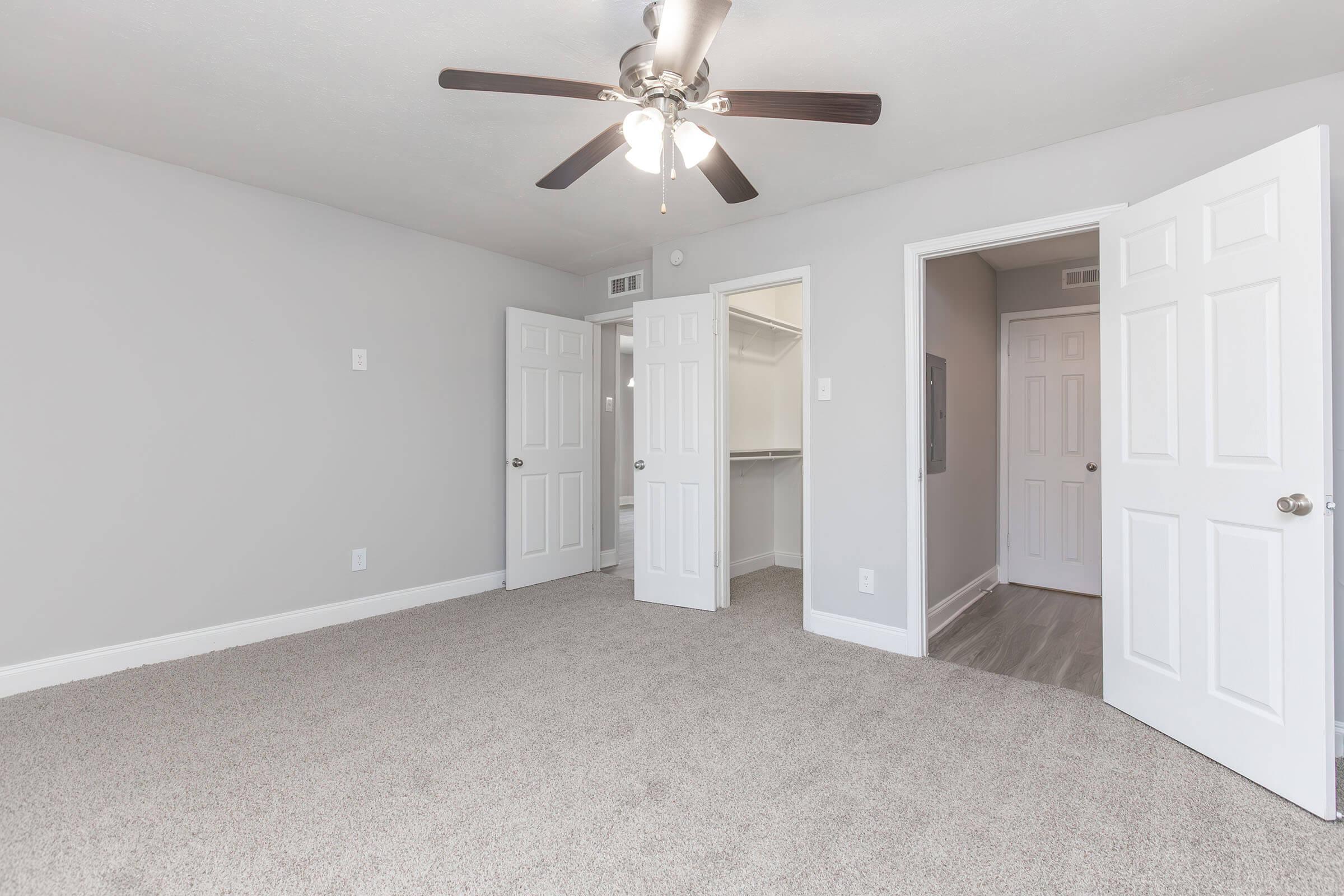 A well-lit, empty bedroom featuring light gray walls, plush carpet, and a ceiling fan. There are two closed doors, one leading to a closet and another to a hallway, with ample space for furniture placement. Natural light enhances the open and airy feel of the room.