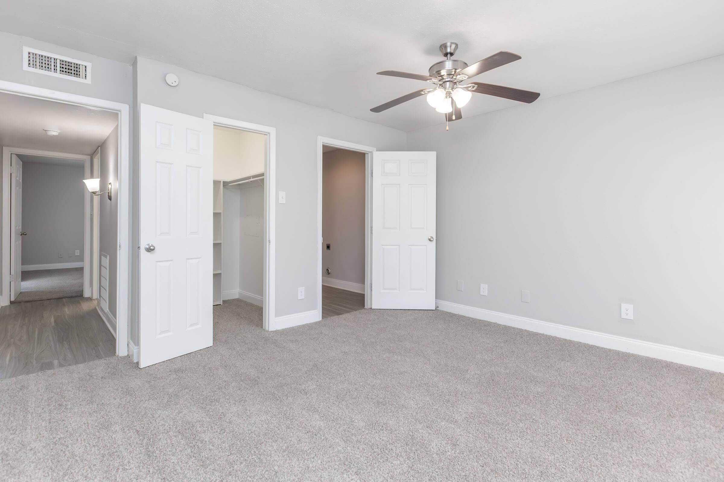 A spacious, light gray bedroom featuring beige carpeting, a ceiling fan, and white doors. The room is well-lit and has two doorways leading to other spaces, with a closet visible on the left. The walls are simple and modern, creating a clean and inviting atmosphere.