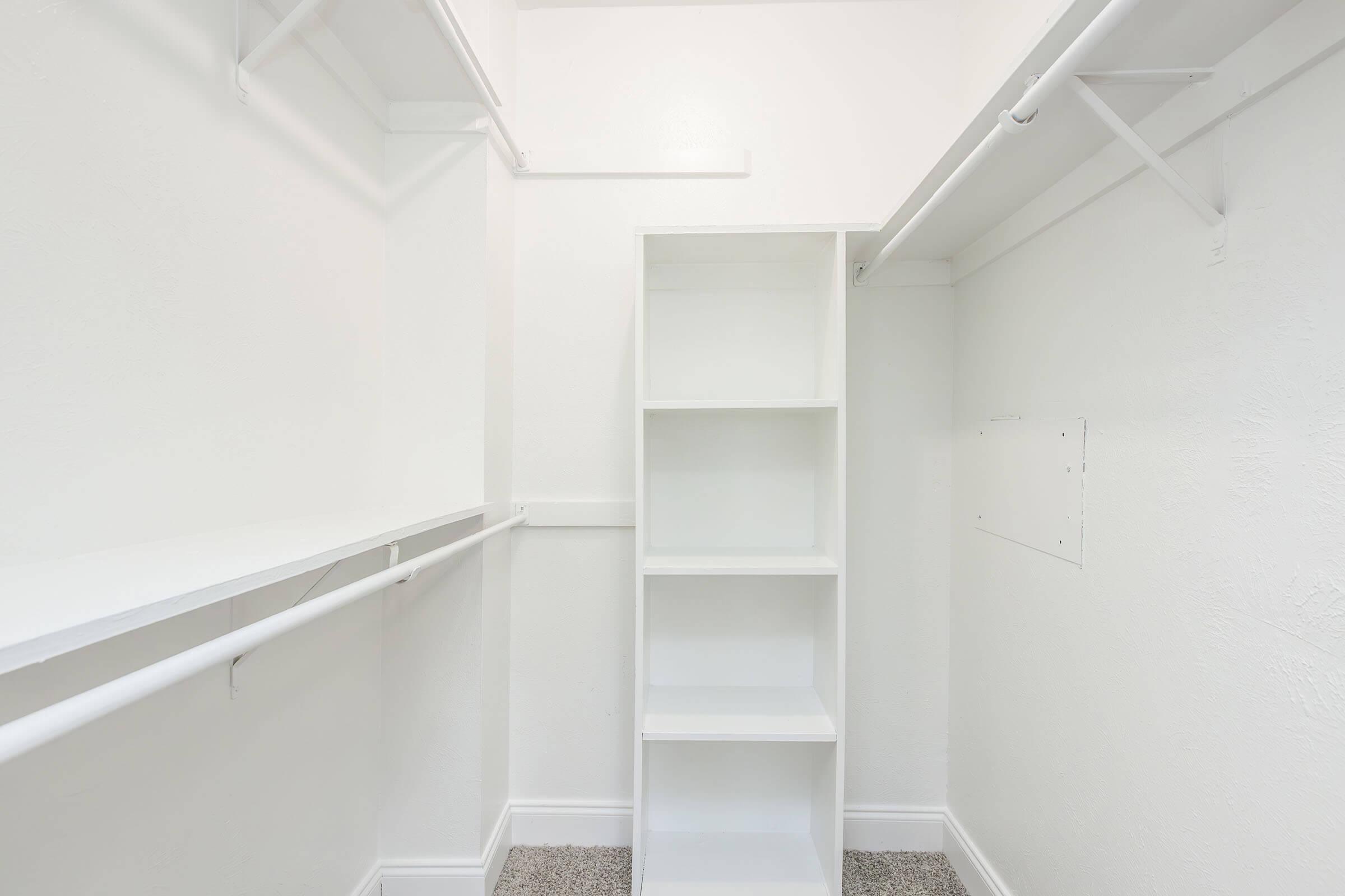 A clean, empty closet with white walls and a light gray carpet. It features two hanging rods on the sides and a tall, narrow white shelving unit in the center, providing storage space. The overall appearance is bright and minimalist, indicating potential for organization and decoration.