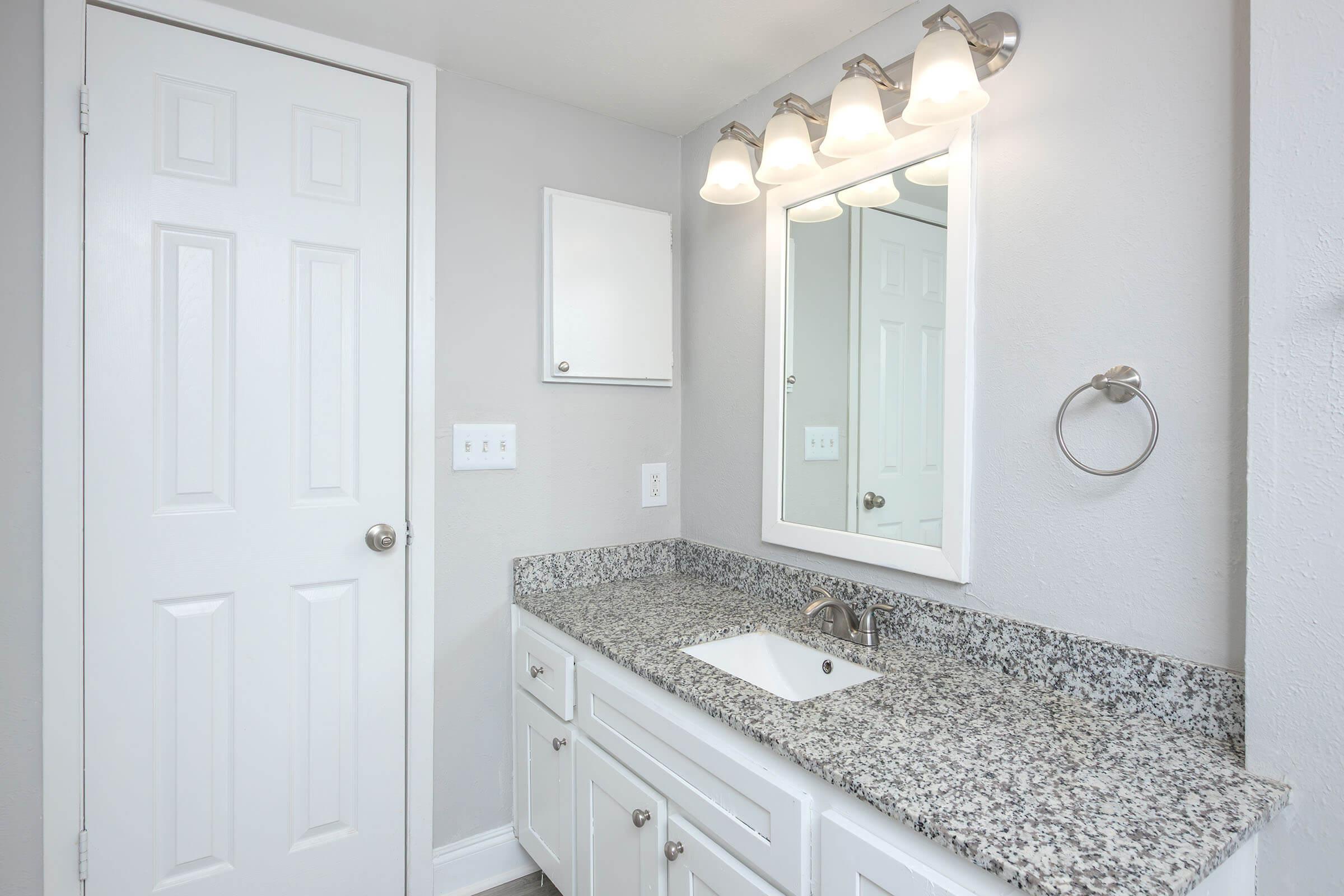 A modern bathroom featuring a granite countertop with a white sink and faucet. Above the sink, there's a rectangular mirror framed in white. The walls are painted light gray, and there's a white door and a light fixture with four bulbs above the mirror, providing ample lighting.
