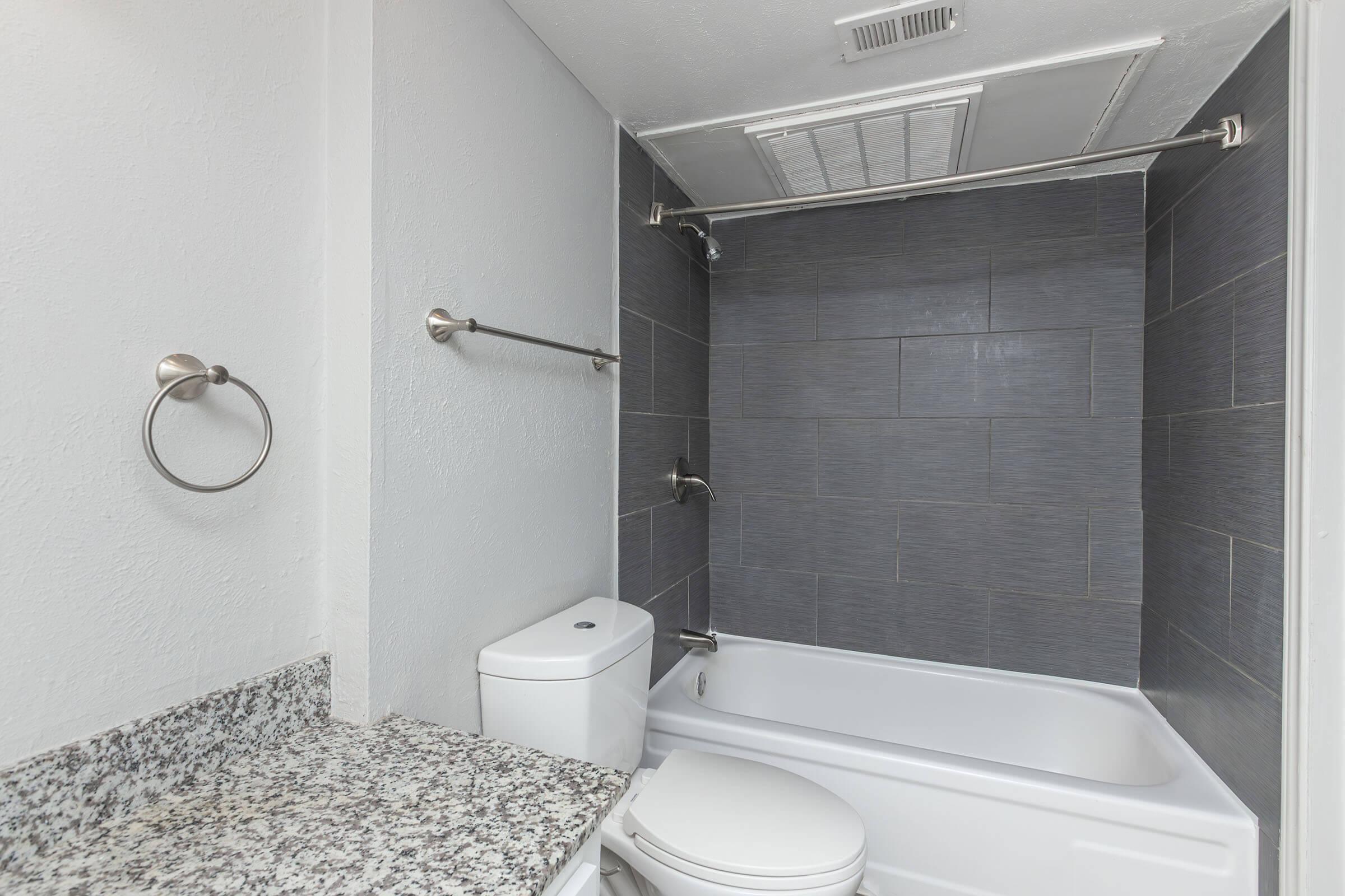 A modern bathroom featuring a white bathtub with a shower curtain rod, gray tiled walls, and a granite countertop. There is a toilet next to the bathtub, and chrome towel racks on the wall, creating a clean and contemporary space.