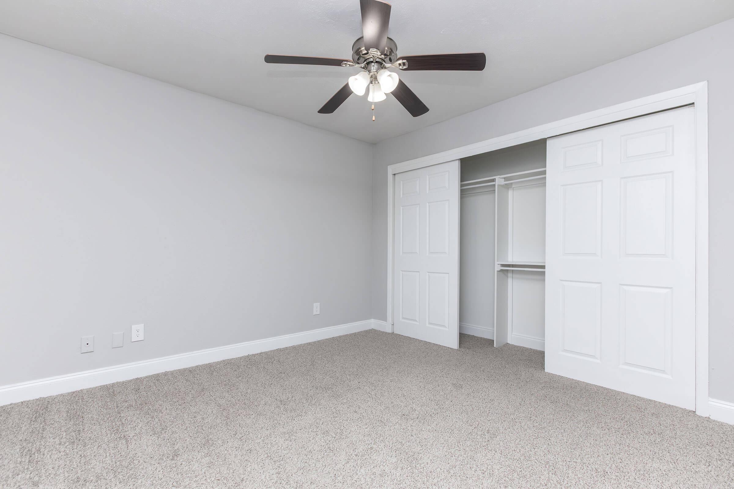 Empty bedroom with light gray walls and plush carpet. A ceiling fan with five blades hangs from the ceiling. There is a double door closet on the right side, featuring white doors that are slightly ajar. The room is well-lit and spacious, providing a minimalist aesthetic.