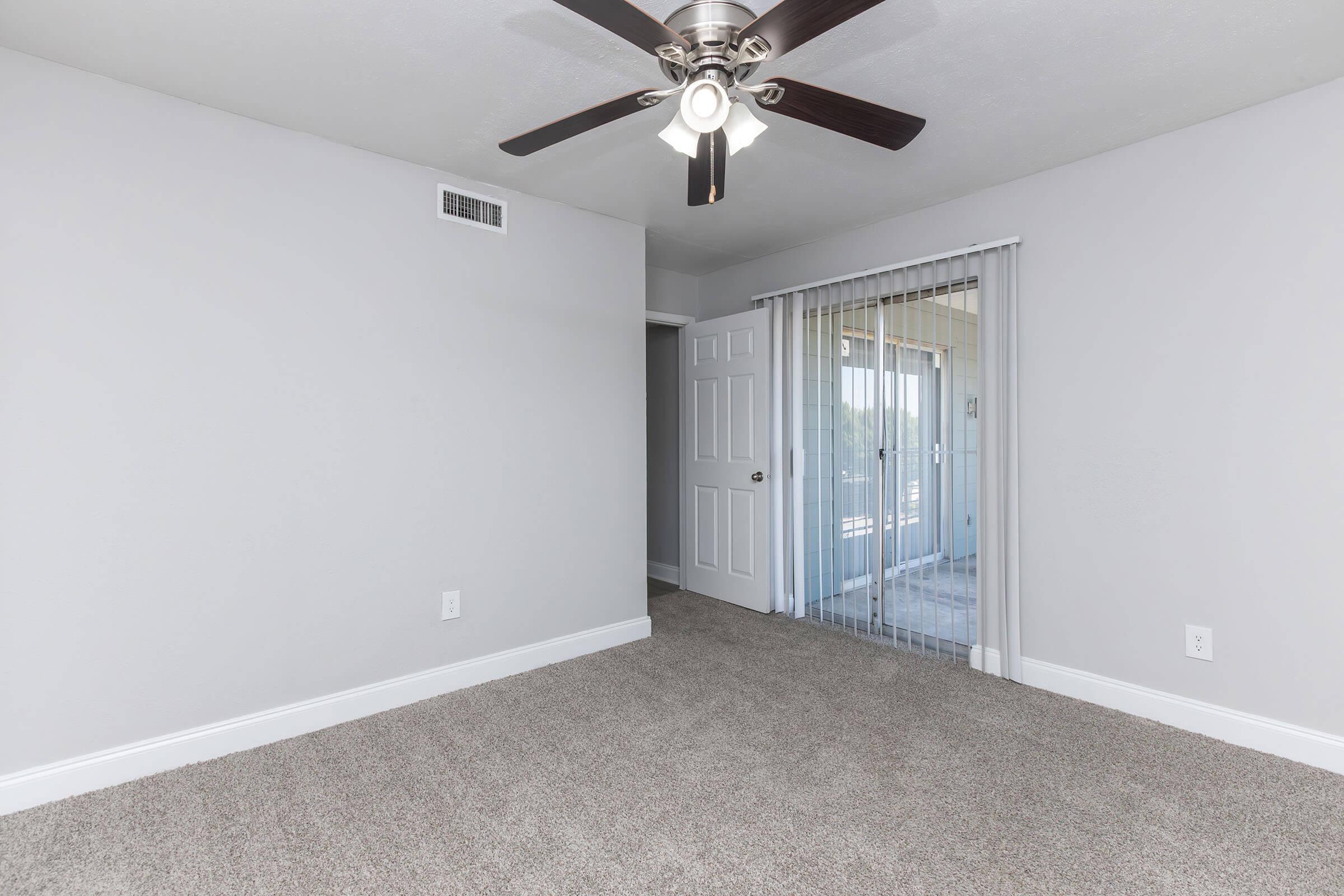 A well-lit, empty room with light gray walls, carpeted flooring, and a ceiling fan. On one side, there is a set of sliding glass doors leading to a balcony or patio, creating a bright and open atmosphere. The room is clean and modern, suitable for various uses.