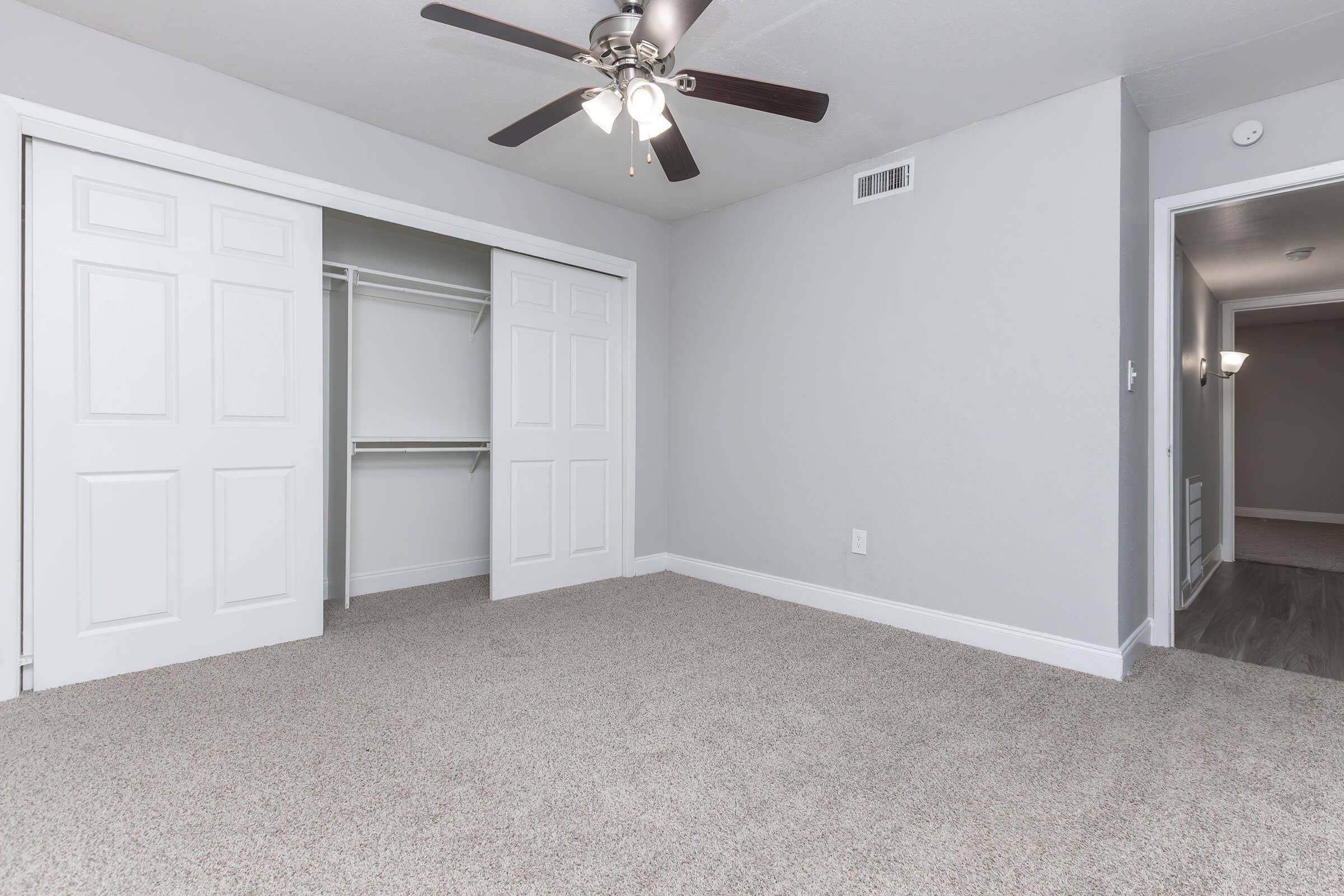 A spacious, empty bedroom featuring light gray walls, a ceiling fan with five blades, and a plush carpeted floor. There are two white double doors leading to a closet on the left and an open doorway on the right, indicating access to another room or hallway. Soft artificial lighting enhances the airy feel of the space.