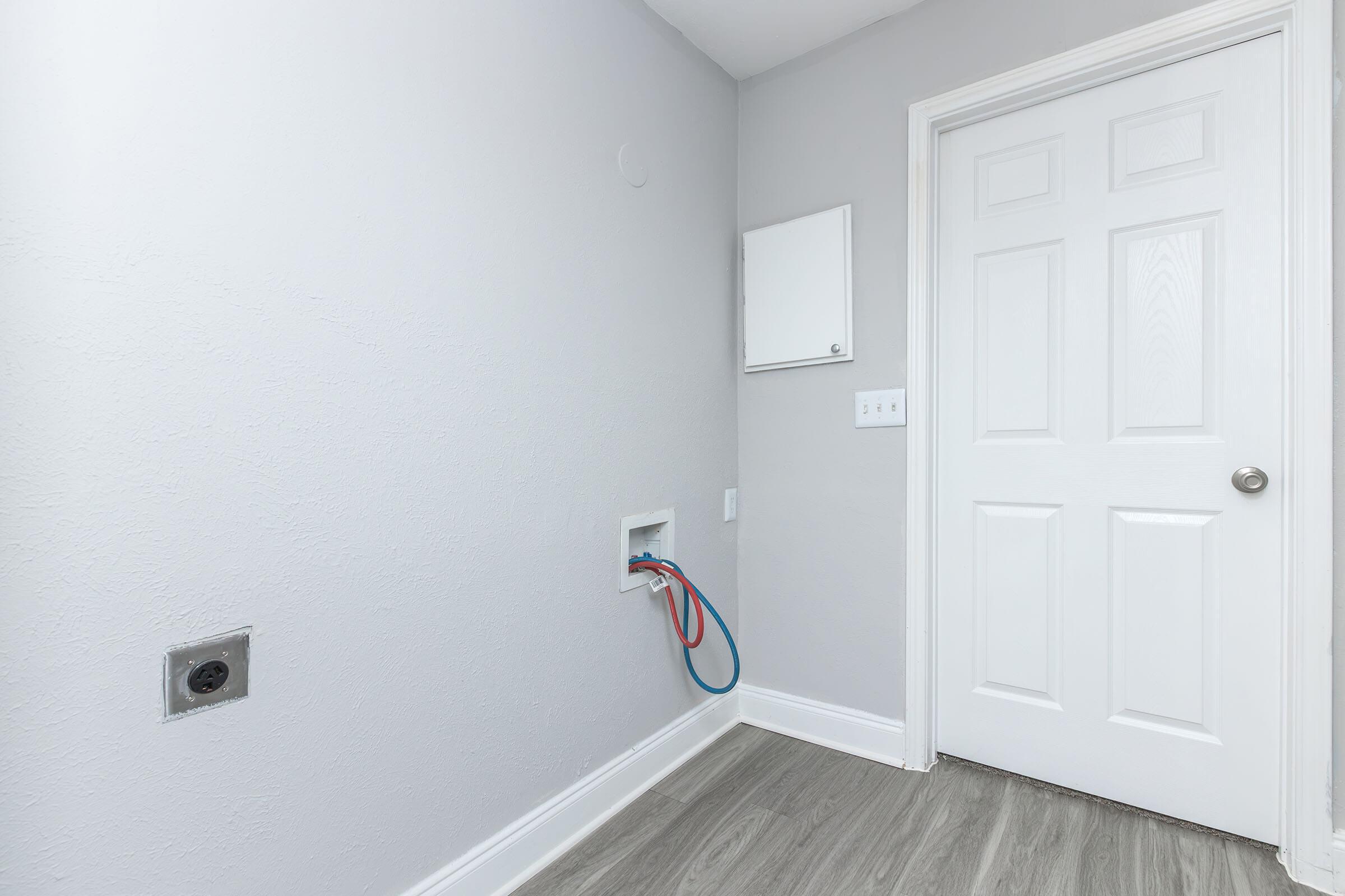 A simple laundry room corner featuring a light gray wall, a closed white door, and a wall-mounted electrical panel. There’s a plumbing connection on the wall with red and blue hoses, but no appliances or furniture in the space. The flooring is a light-toned wood laminate.