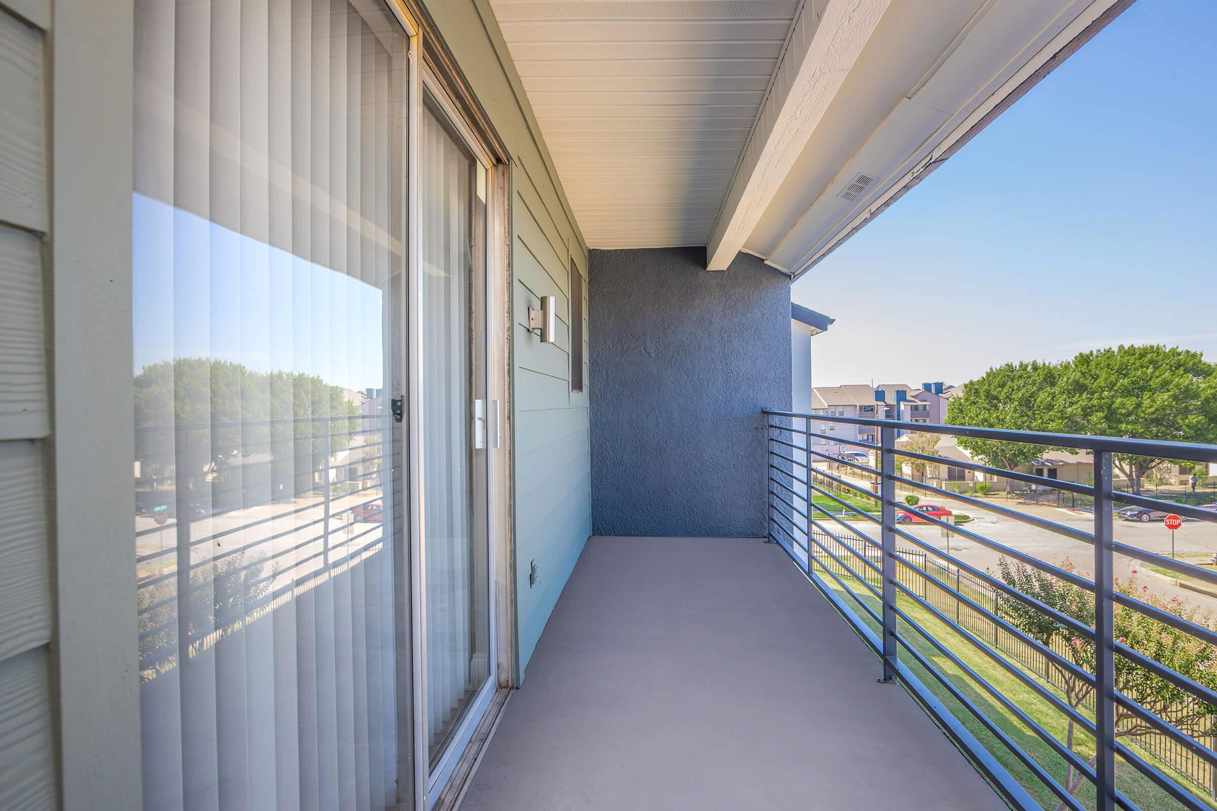 A view of a balcony with glass sliding doors, featuring a railing and a clear sky in the background. The balcony is empty, with light-colored walls and a concrete floor. Green trees and a street are visible in the distance.