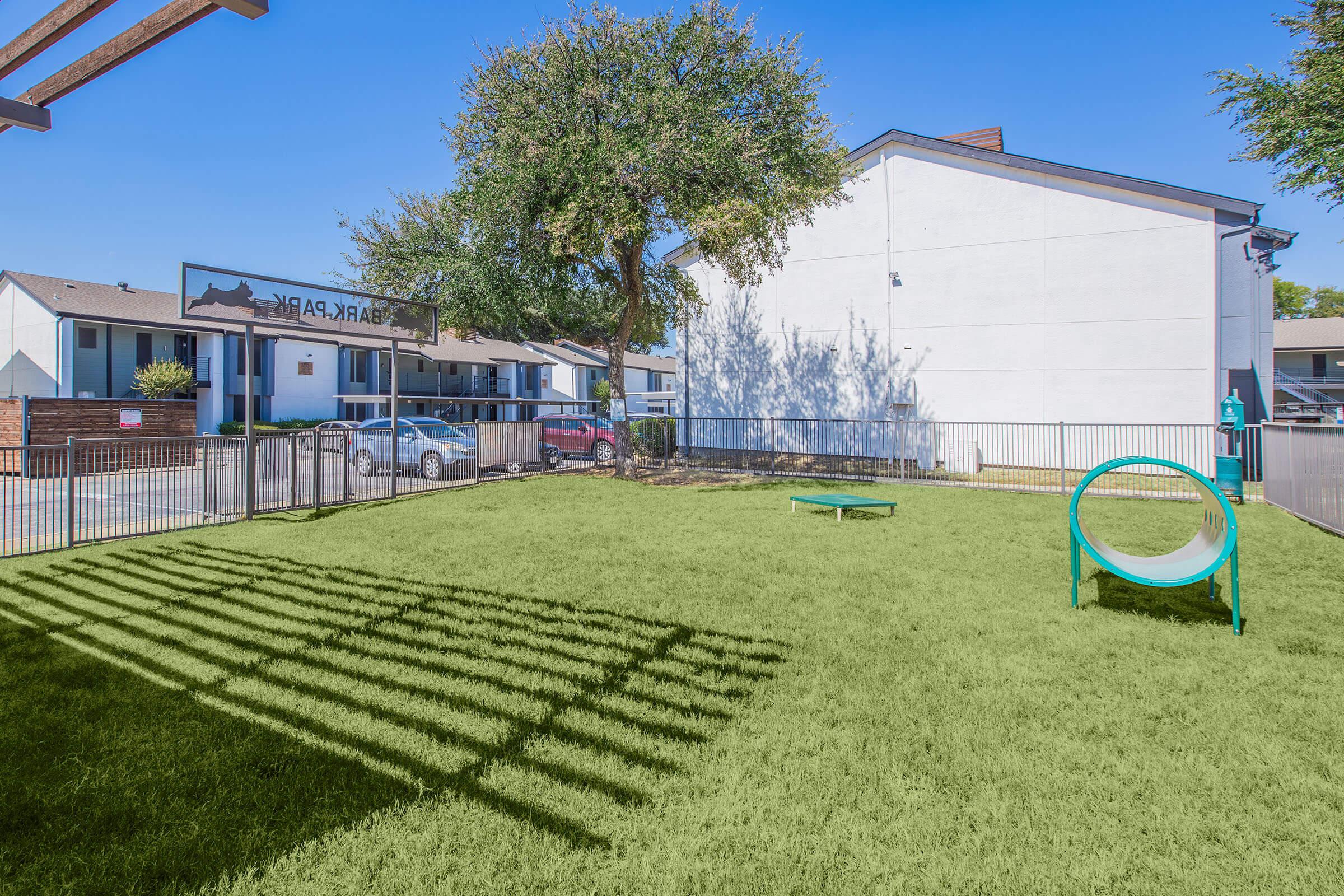 A sunny outdoor park area featuring green grass, a tunnel for pets, and a fence. In the background, there are buildings with an inviting atmosphere, and a sign indicating the area is called "Bark Park." The clear blue sky enhances the lively setting.