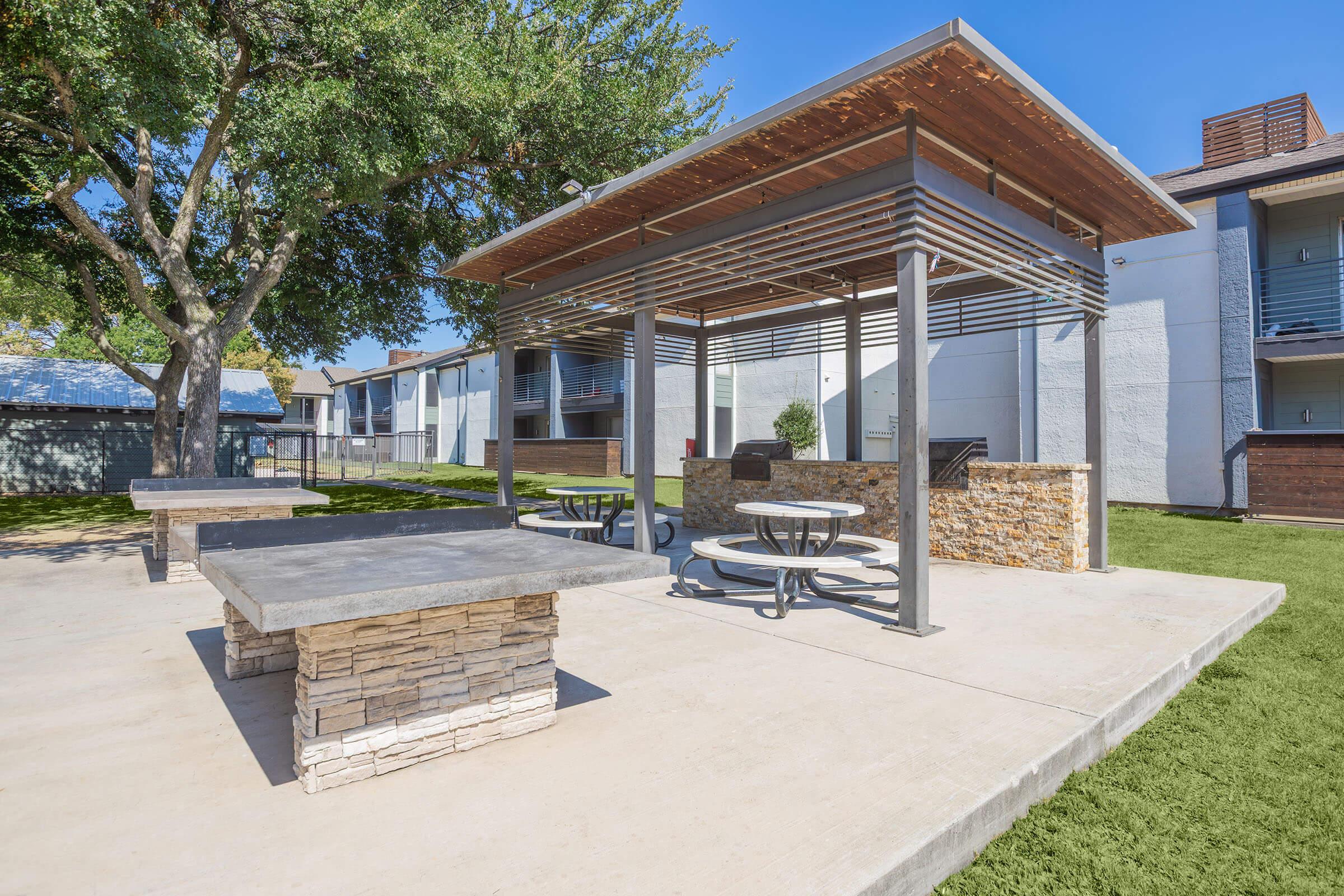 A landscaped outdoor area featuring a stone and metal pavilion with picnic tables. Surrounding greenery includes grass and trees. In the background, there are apartment buildings with balconies under a clear blue sky.