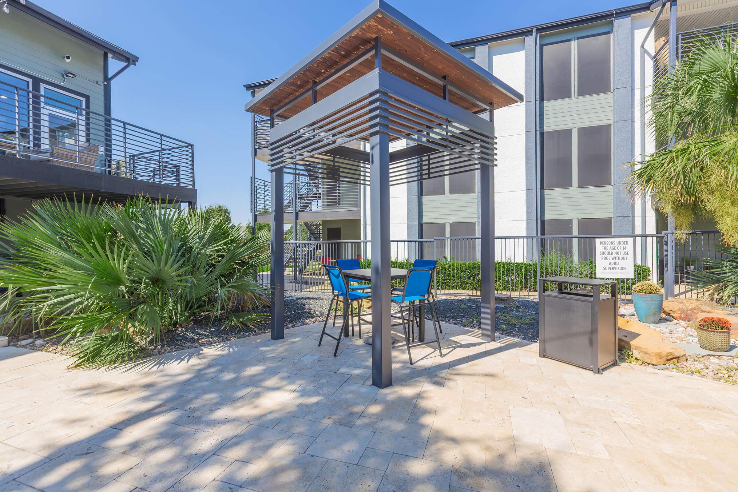A sunlit outdoor seating area featuring a metal gazebo with two blue chairs and a table, surrounded by palm plants. In the background, there are modern apartment buildings. The scene is calm and inviting, ideal for relaxation or socializing.