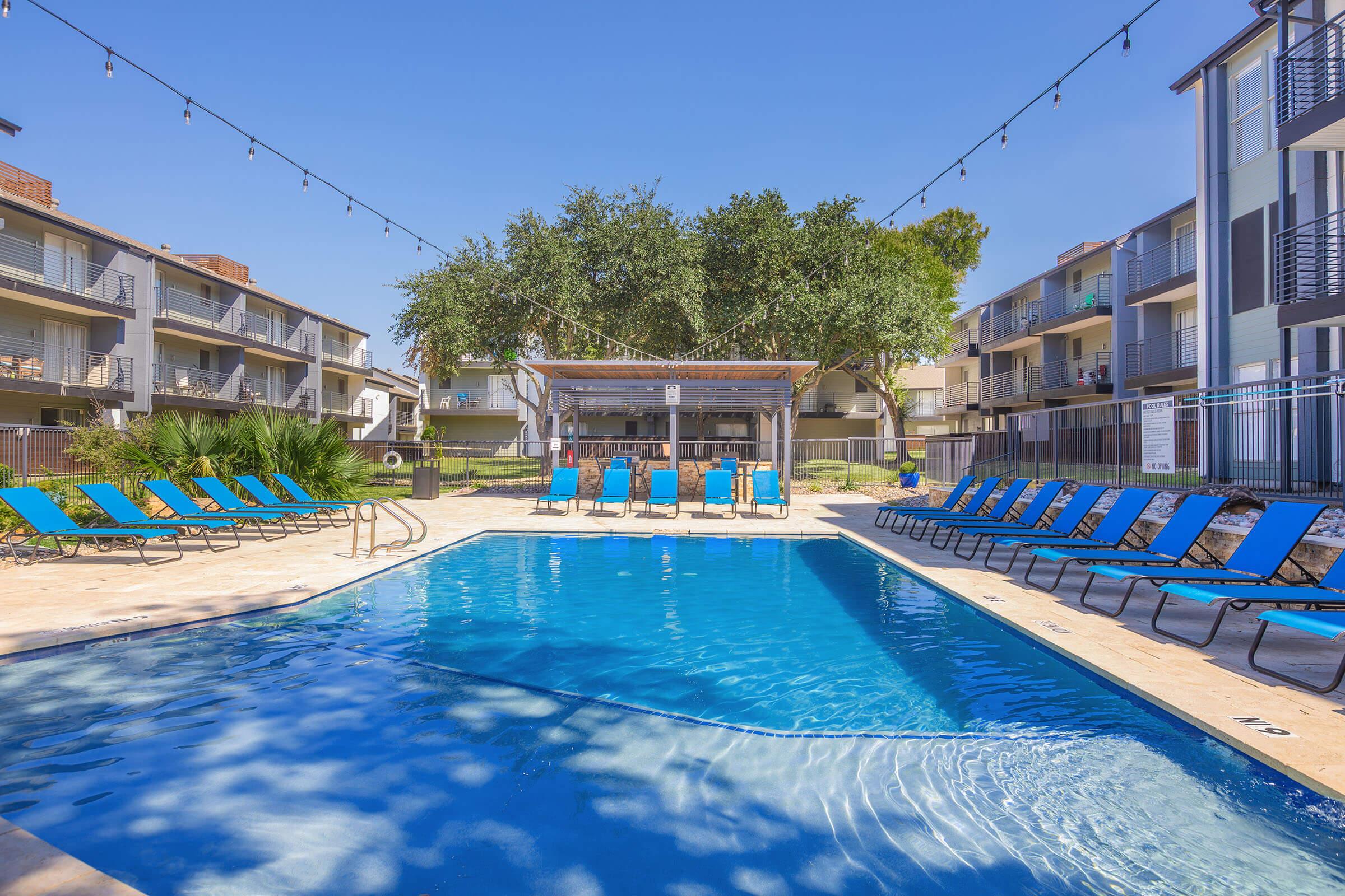 A swimming pool surrounded by blue lounge chairs, with a shaded cabana area in the background. Tall trees provide some greenery, and apartment buildings are visible on either side of the pool area. String lights are hung above, creating a relaxed outdoor atmosphere.