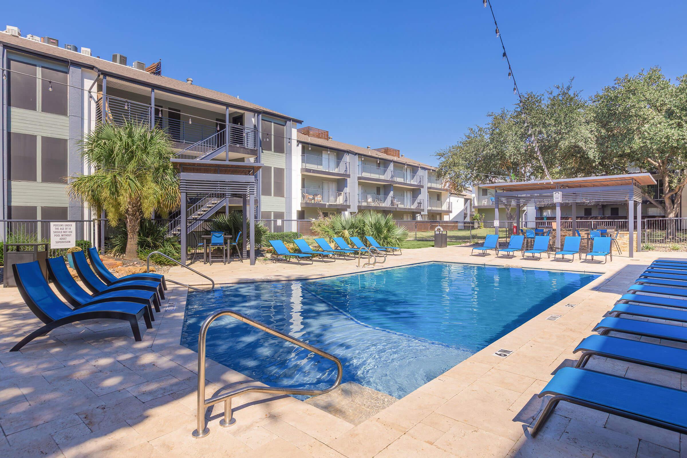 A sunny pool area featuring a clear blue swimming pool, surrounded by lounge chairs and palm trees. Multi-story buildings can be seen in the background, with some outdoor seating areas. String lights are hung above the pool for ambiance.