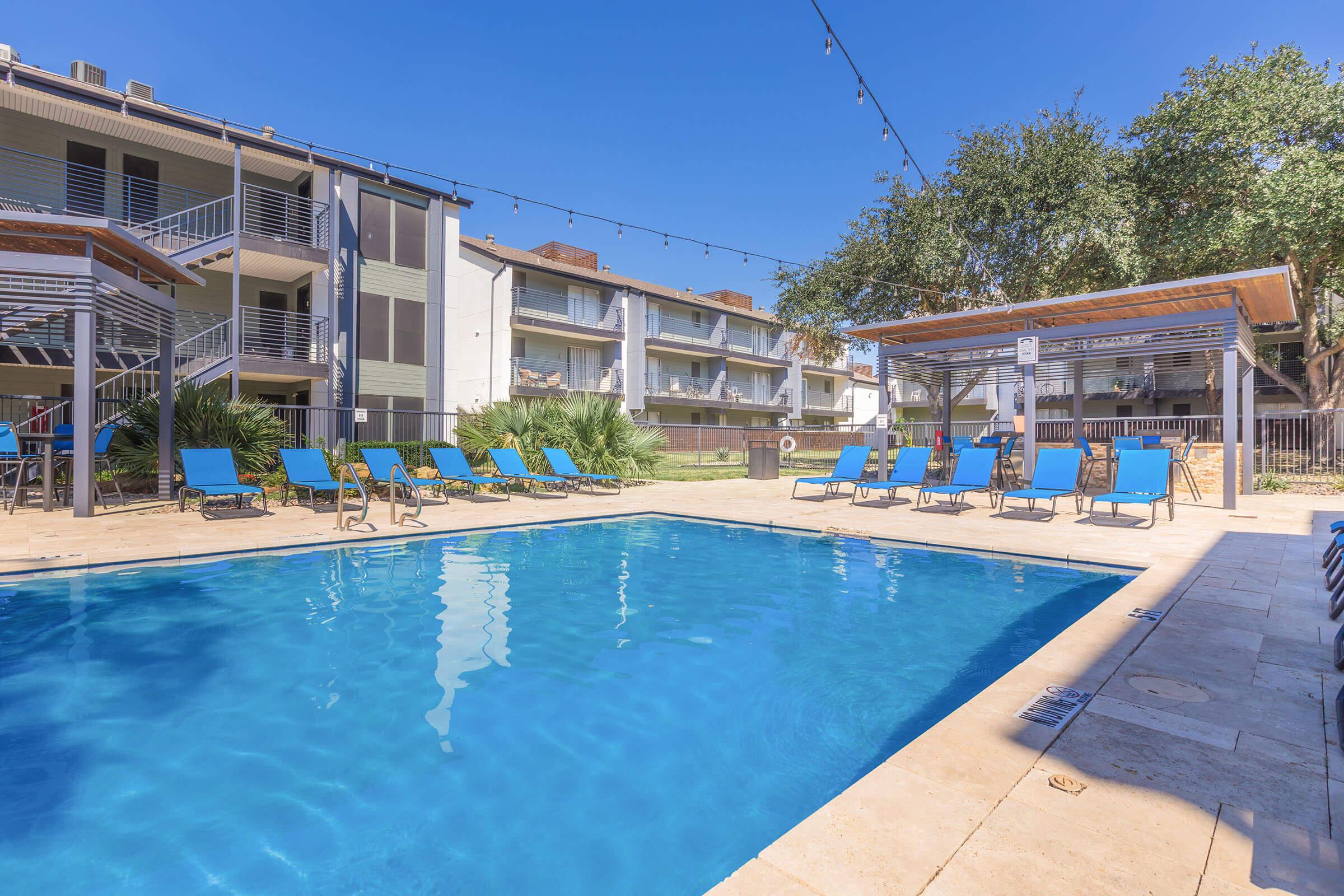 A clear blue swimming pool surrounded by lounge chairs under sunny skies. There are two cabanas on either side of the pool, and apartment buildings are visible in the background. Decorative string lights hang above, creating a relaxed atmosphere. Palm trees add greenery to the scene.