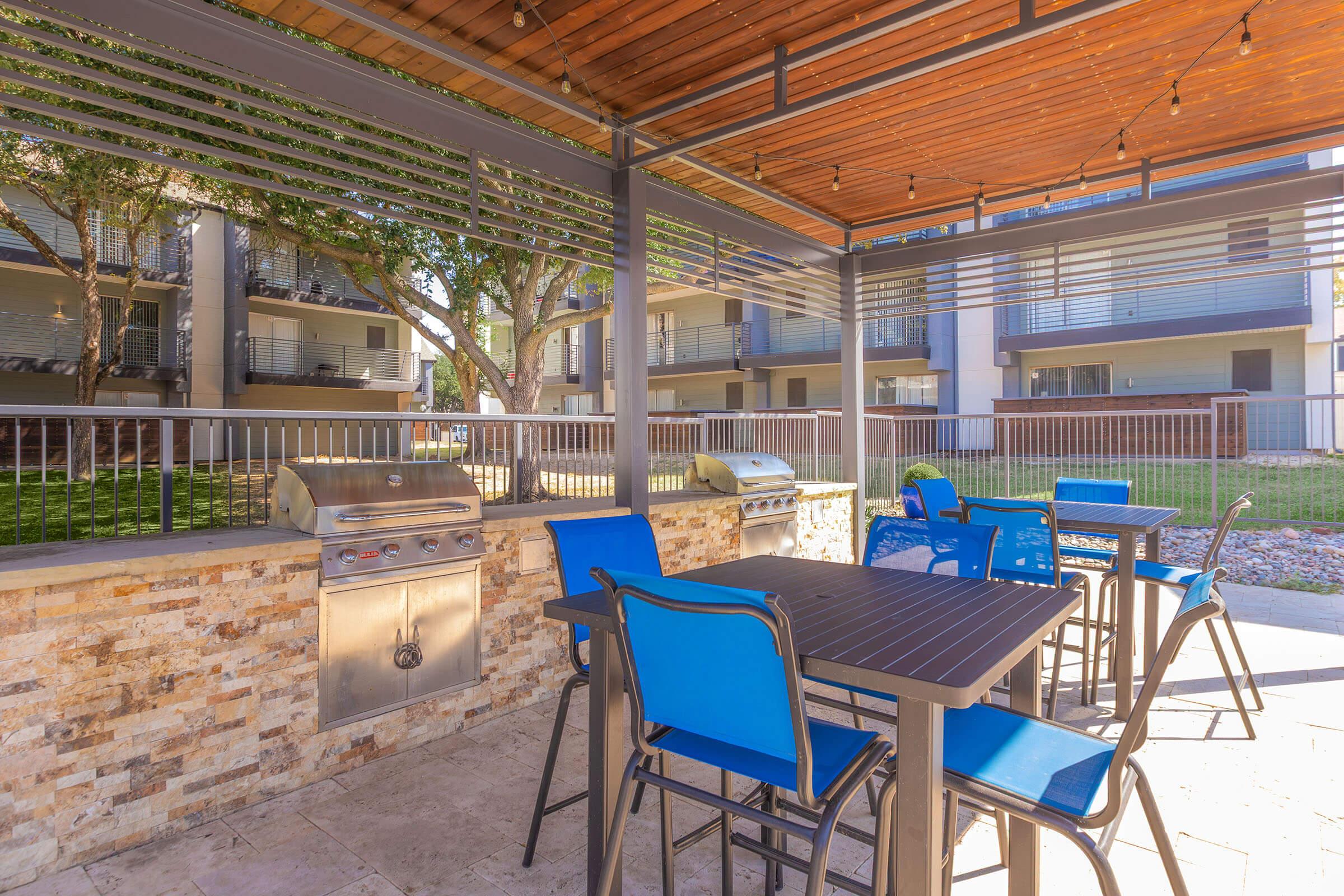 An outdoor grilling area featuring a stainless steel barbecue grill, a stone countertop, and several blue chairs around a dark table. The setting is shaded, with nearby greenery and a view of apartment buildings in the background, creating a relaxed atmosphere for social gatherings.