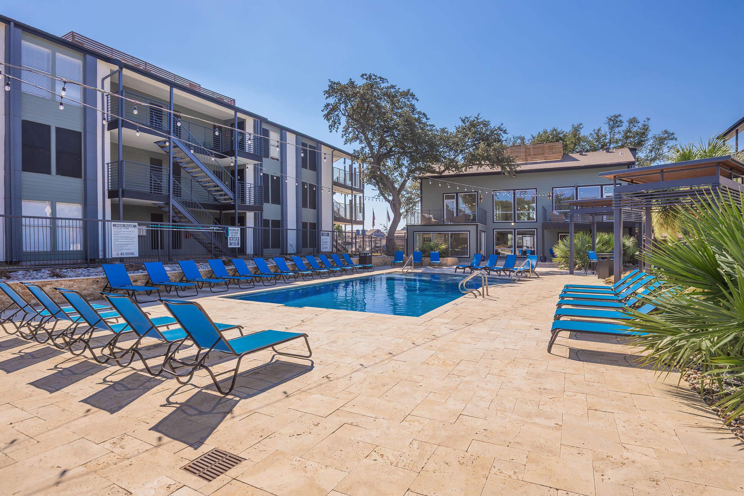 A sparkling outdoor swimming pool surrounded by lounge chairs, with a modern building in the background. The area is sunny and inviting, featuring landscaped greenery and a clear blue sky. Stairs lead to upper-level apartments on one side, creating a resort-like atmosphere.