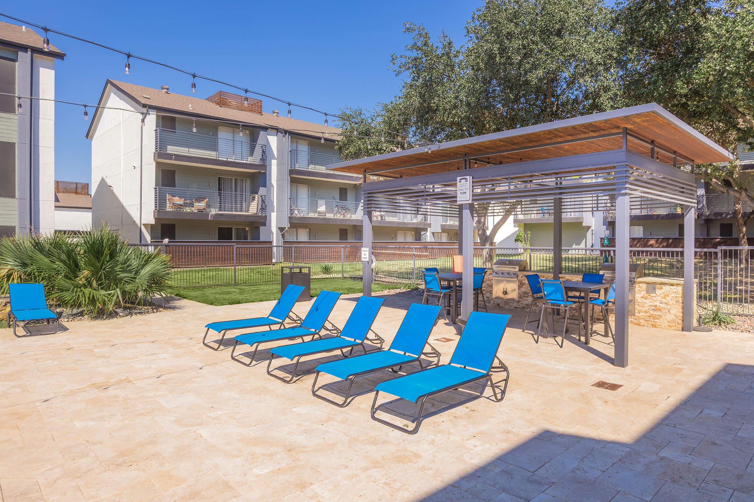A sunny outdoor area featuring several blue lounge chairs arranged around a stone patio. In the background, there are multi-story apartment buildings with balconies. A shaded gazebo with seating is visible nearby, along with landscaped greenery. The scene conveys a relaxed, inviting atmosphere.