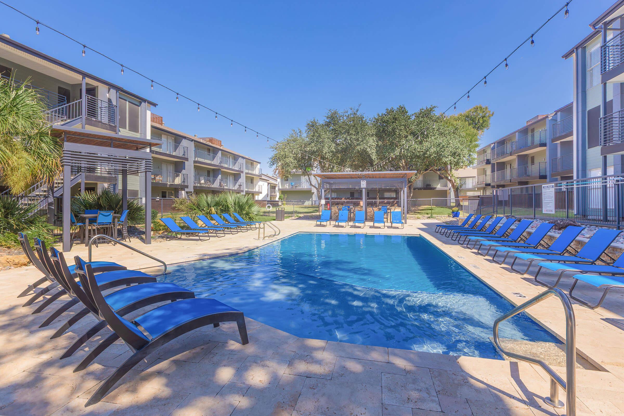 A serene outdoor pool surrounded by lounge chairs, with a few shaded areas and palm trees in the background. The sky is clear and blue, and string lights are hanging above the pool area, creating a relaxing atmosphere. Modern apartment buildings are visible in the background.