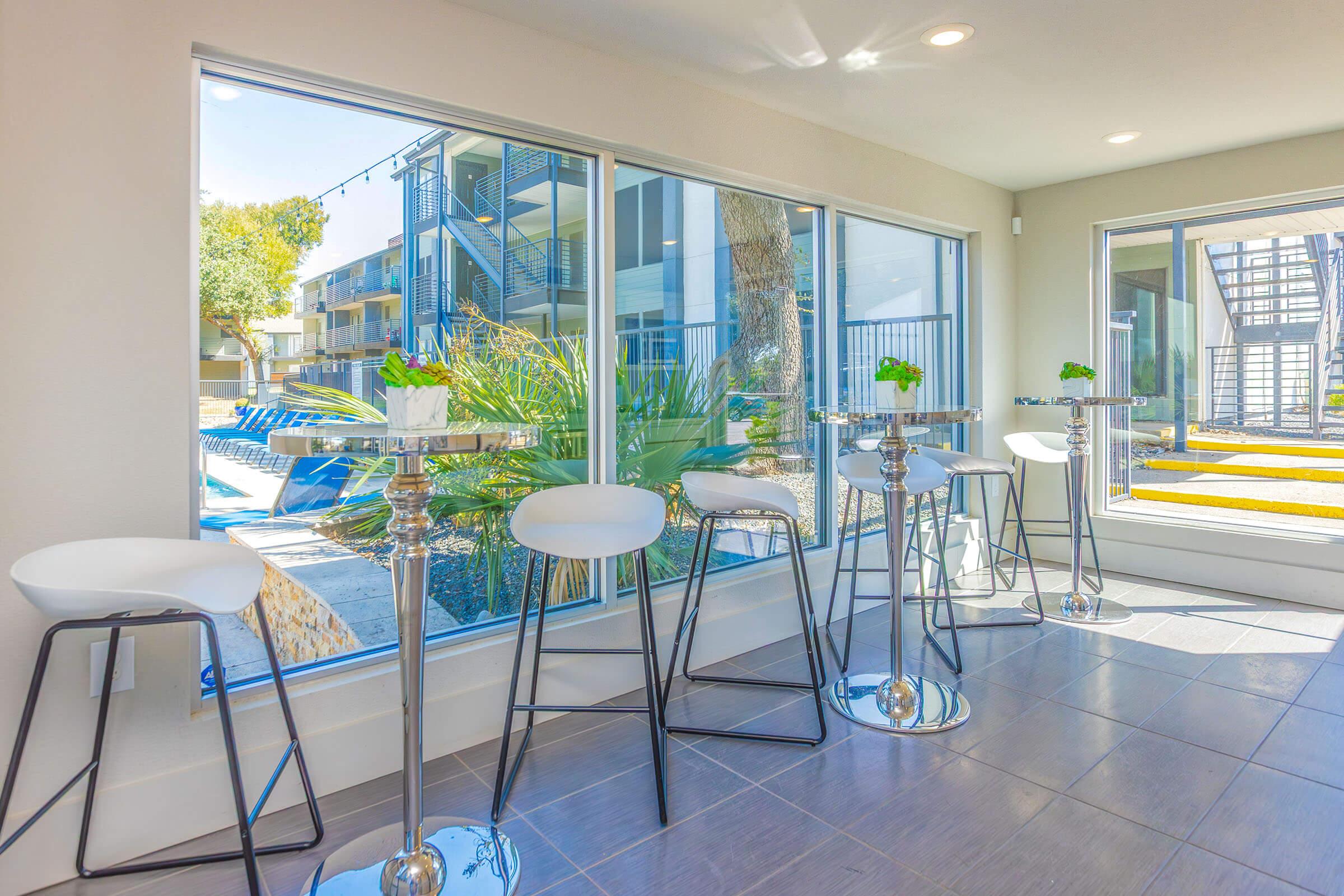 A modern seating area featuring tall, sleek bar stools around small tables, with large windows offering a view of an outdoor pool and greenery. Natural light floods the space, enhancing its contemporary design and inviting atmosphere.
