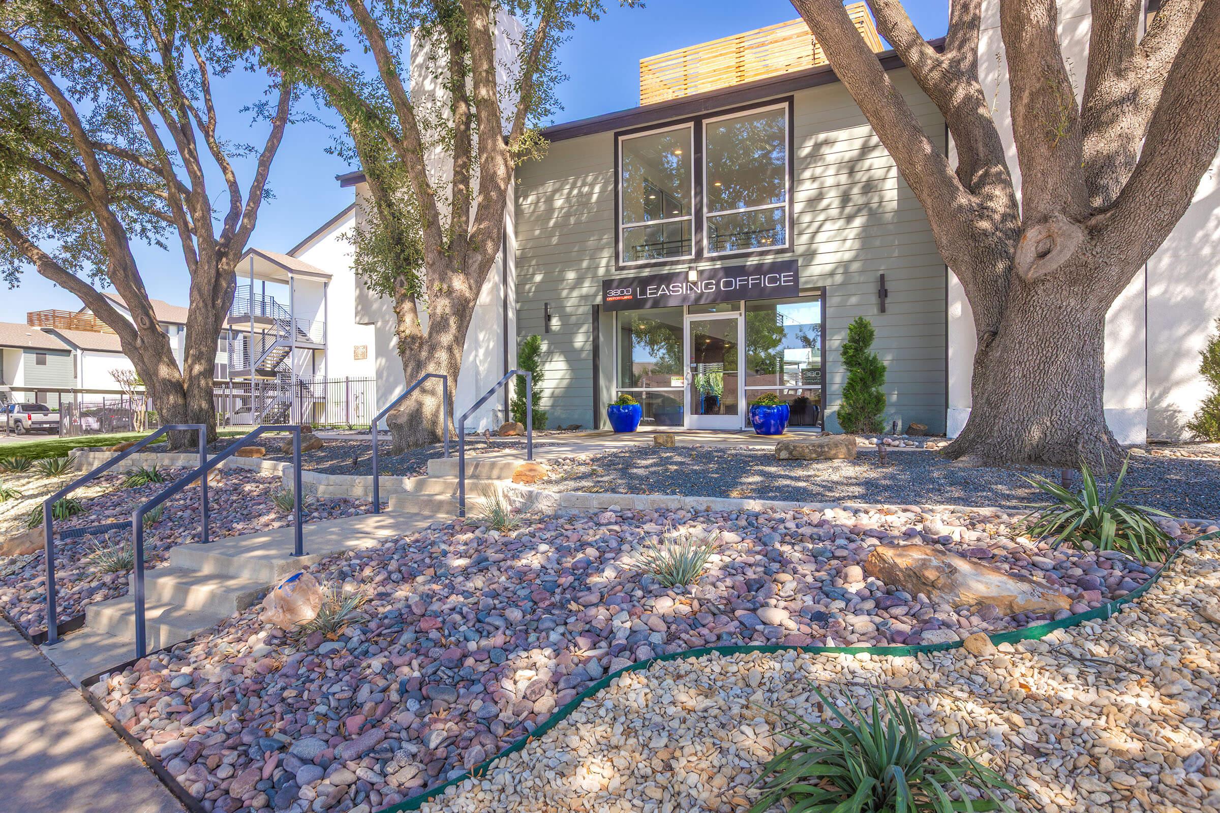 A modern leasing office building with large windows, surrounded by landscaped gardens featuring rocks, plants, and trees. The entrance is marked by steps leading up to the office door, with blue planters on either side, and a sunny blue sky overhead.