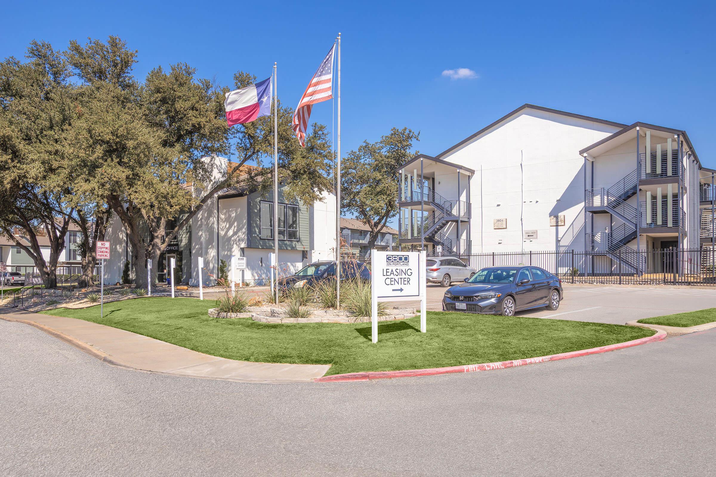 A leasing center for an apartment complex, featuring Texas and American flags in front. The building has a modern design with two stories and a parking area visible. The landscape includes green grass and trees, set against a clear blue sky.