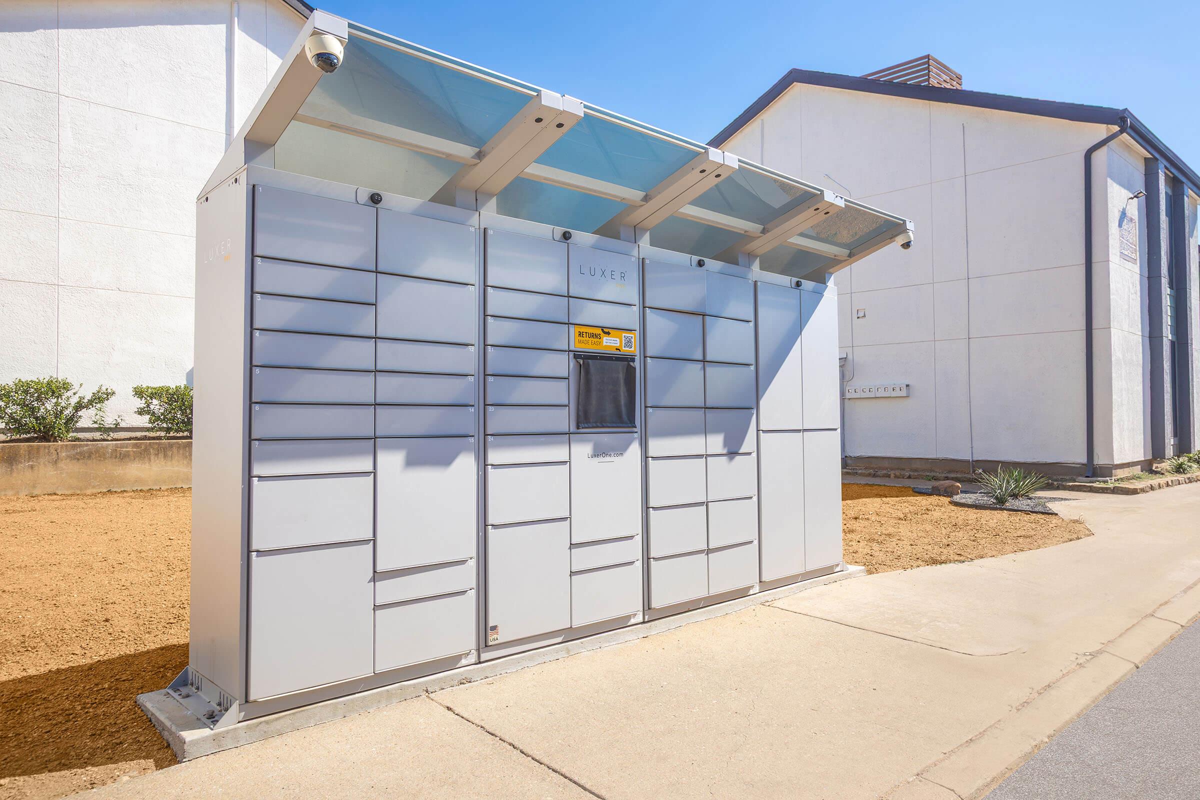 A modern package delivery locker system with multiple compartments, designed for secure retrieval of parcels. The lockers are set against a backdrop of a building and landscaping, featuring a clear blue sky above.