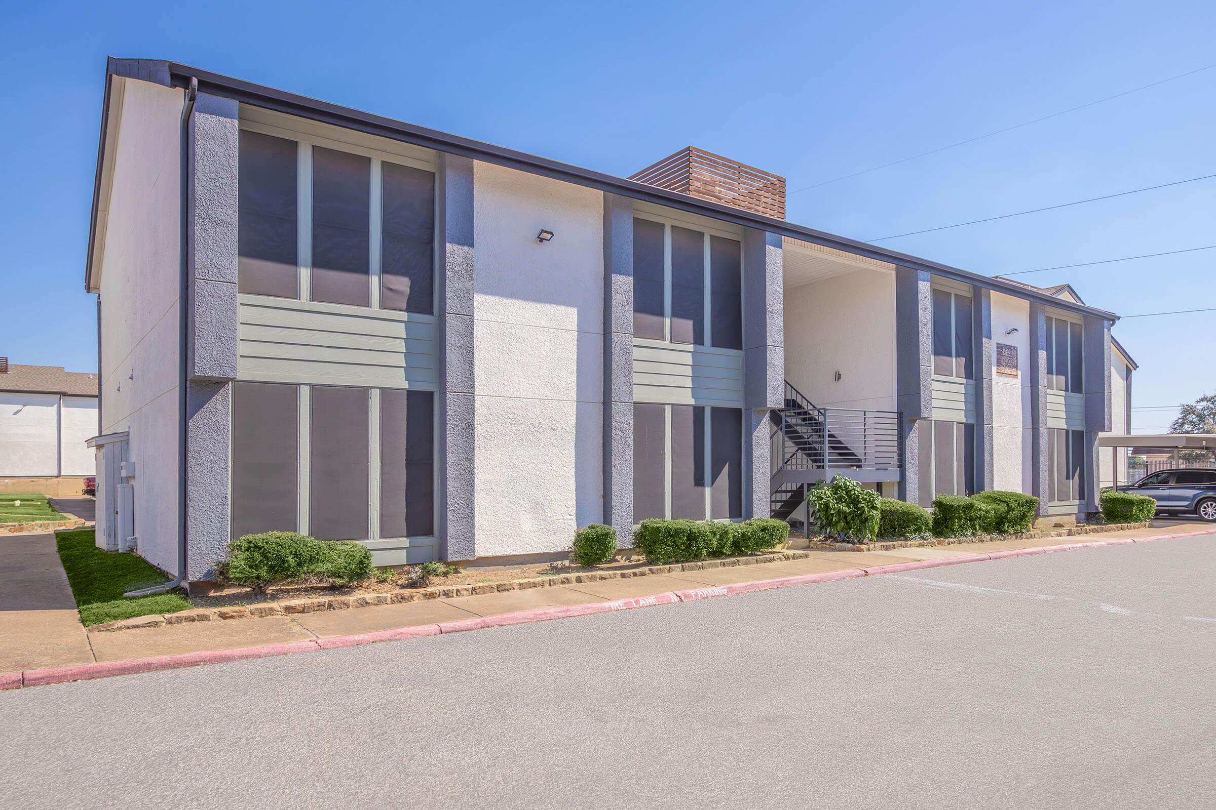 Exterior view of a two-story apartment building featuring a white facade with gray accents, large windows, and a staircase leading to the second floor. Well-maintained landscaping is visible in front, along with a paved parking area. The sky is clear and blue.