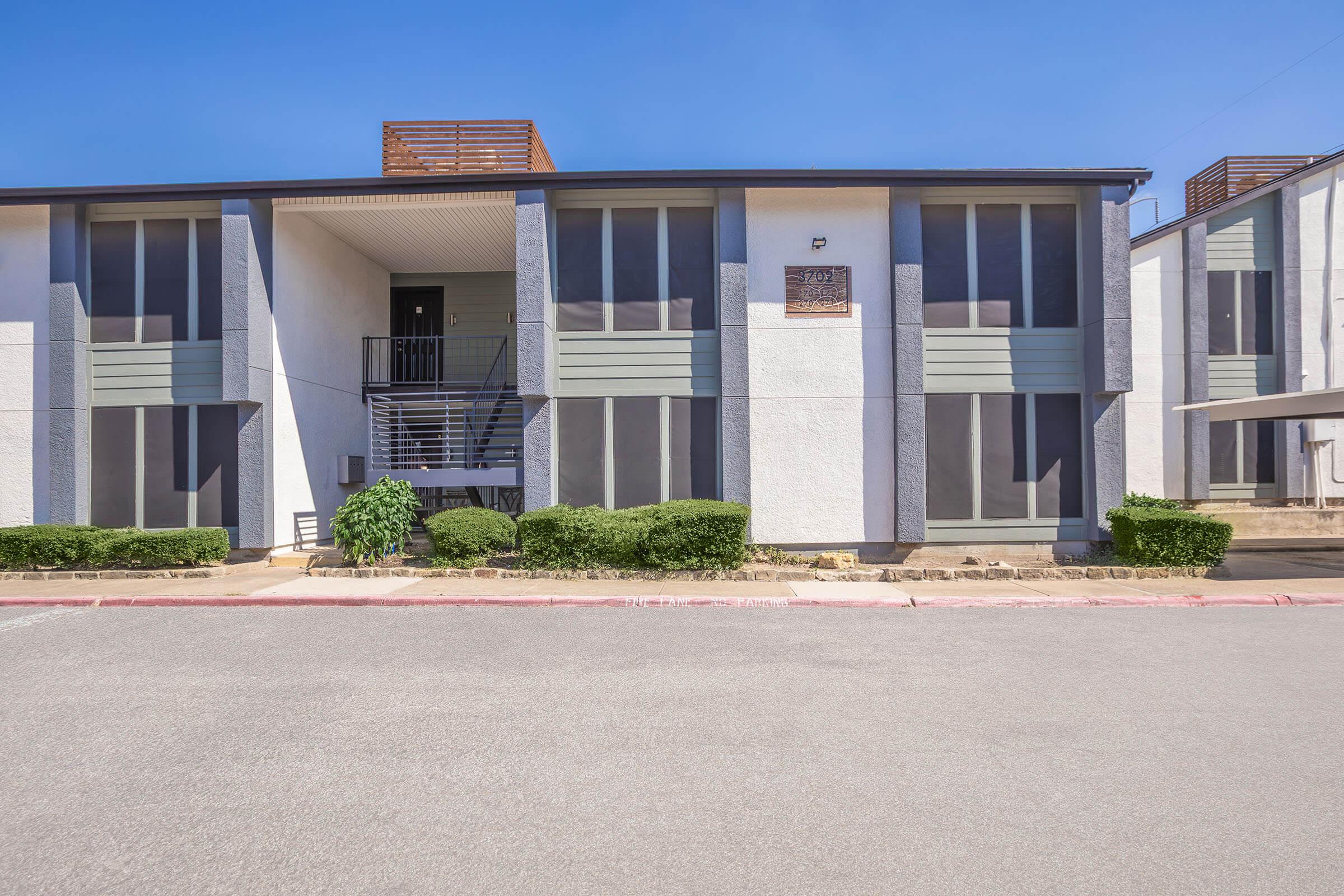Exterior view of a two-story apartment building featuring large windows, a staircase leading to the entrance, and manicured shrubs in front. The building has a modern design with a light-colored facade and is situated on a paved driveway under a clear blue sky.