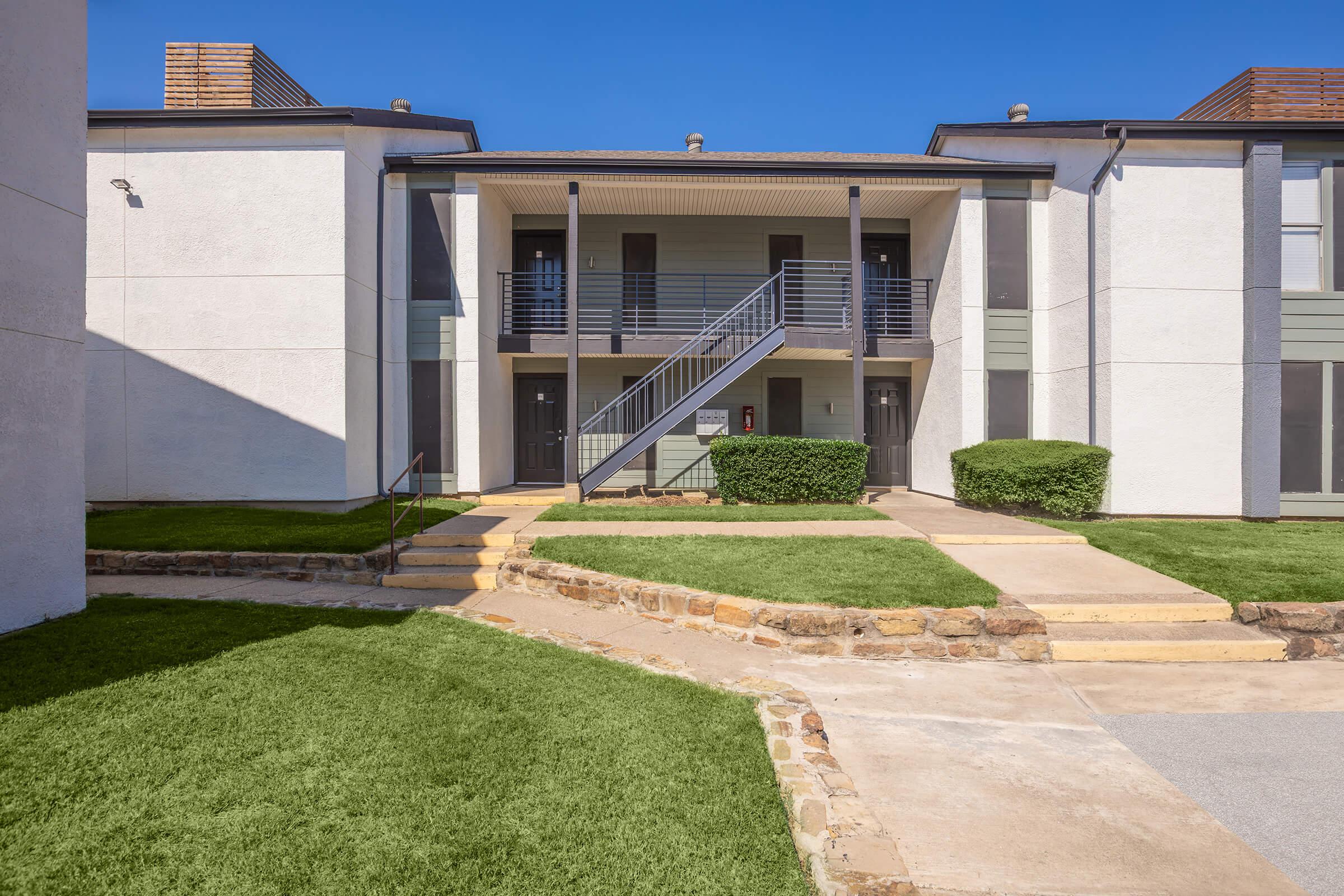 A view of an apartment complex featuring two building structures with a central pathway. A staircase leads to the upper level entrance of one building. The area is landscaped with green grass and small bushes, under a clear blue sky.