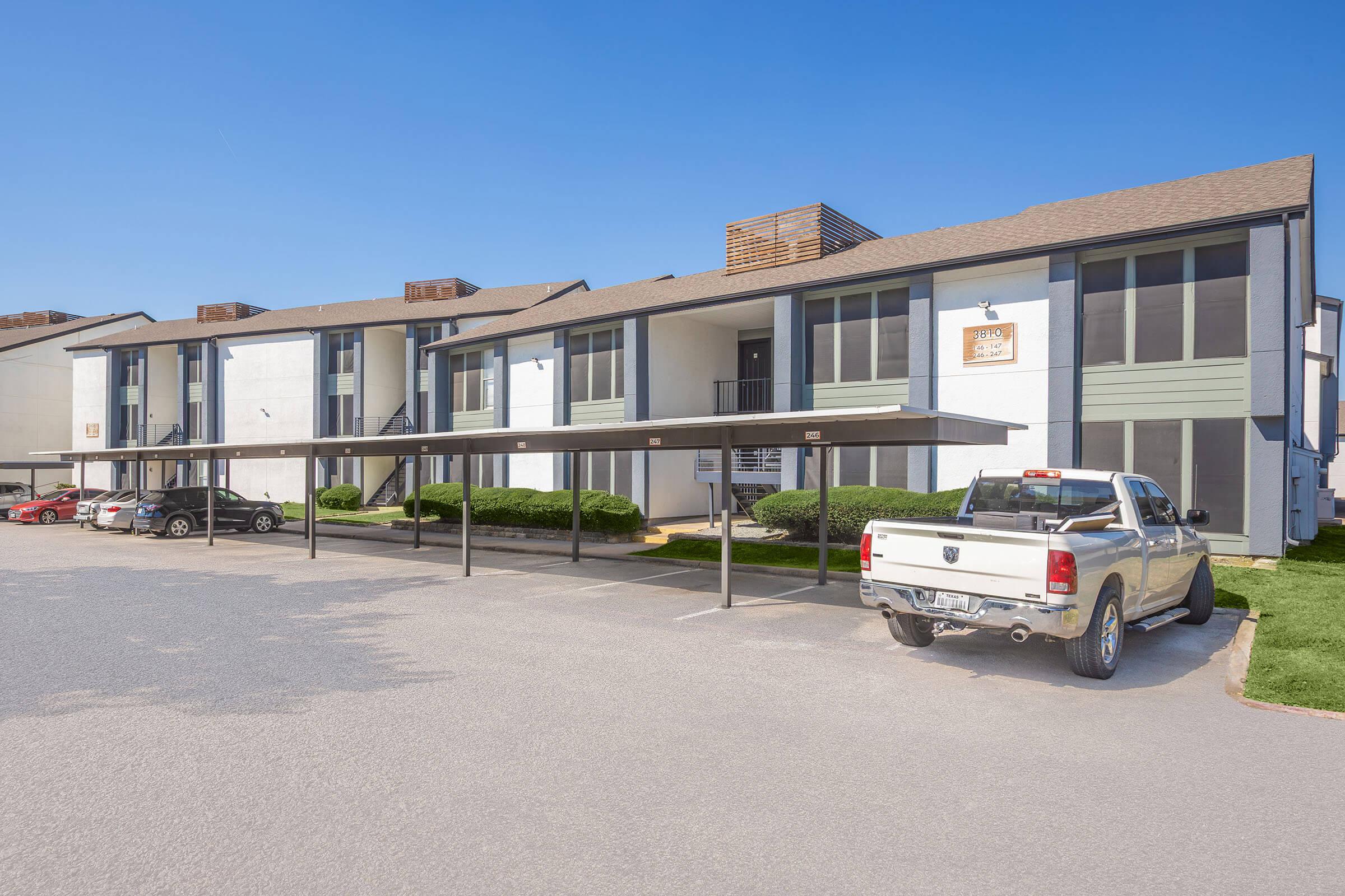 A two-story apartment complex with a light-colored exterior and green accents. Several covered parking spaces are visible in front of the building, along with parked cars. The sky is clear and blue, and neatly trimmed bushes line the walkway to the entrance.