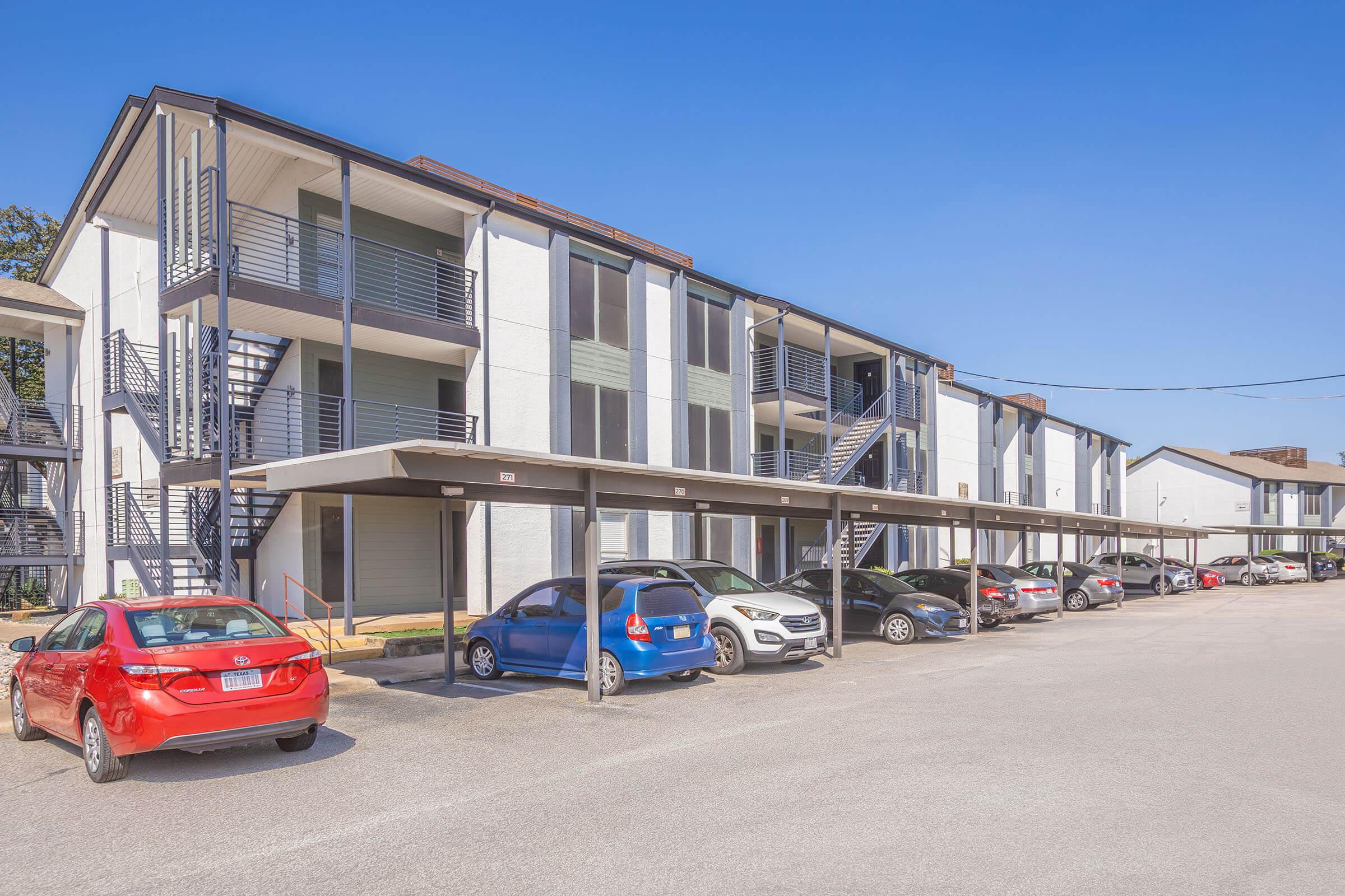 A modern apartment complex with two-story buildings and covered parking. Several cars are parked under awnings in front of the buildings, which feature balconies and staircases. The sky is clear and blue, indicating a sunny day.