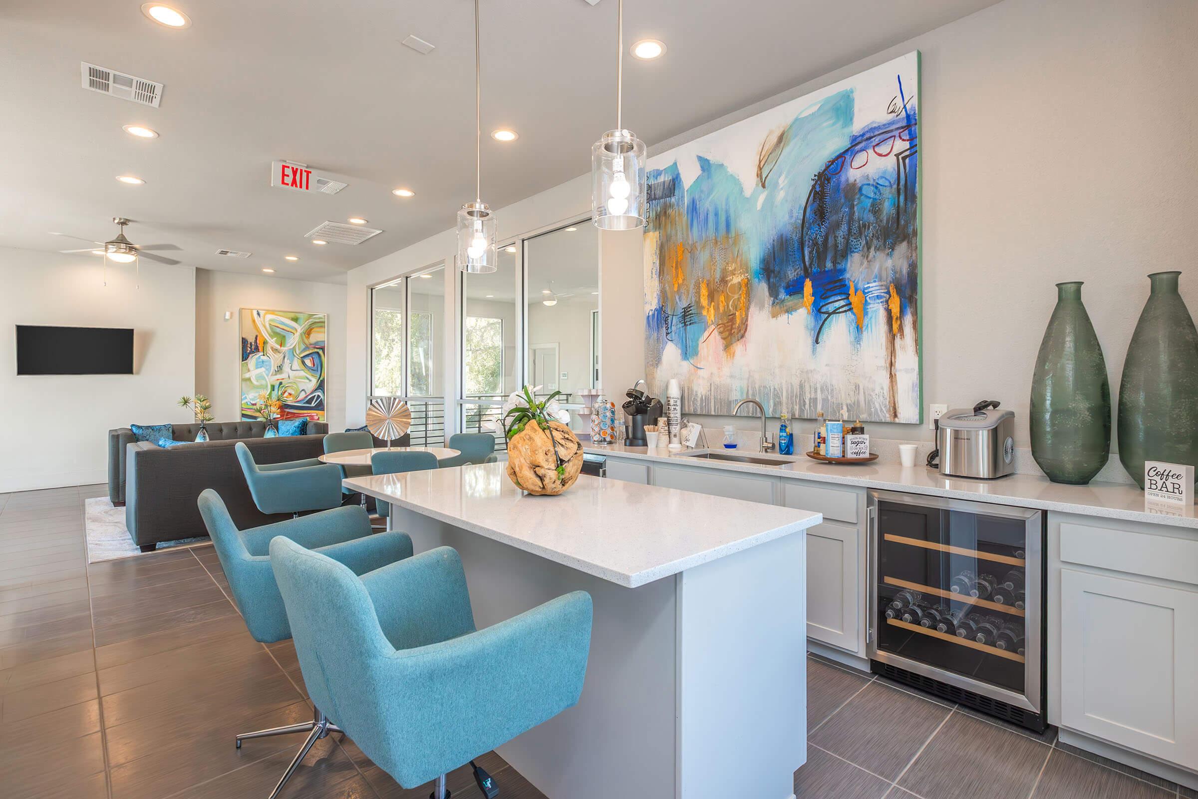 A modern interior of a stylish lounge area featuring a light-colored bar with seating, decorative vases, a wine cooler, and artwork on the wall. The space includes sofas, a television, and large windows allowing natural light to fill the room, creating an inviting atmosphere.
