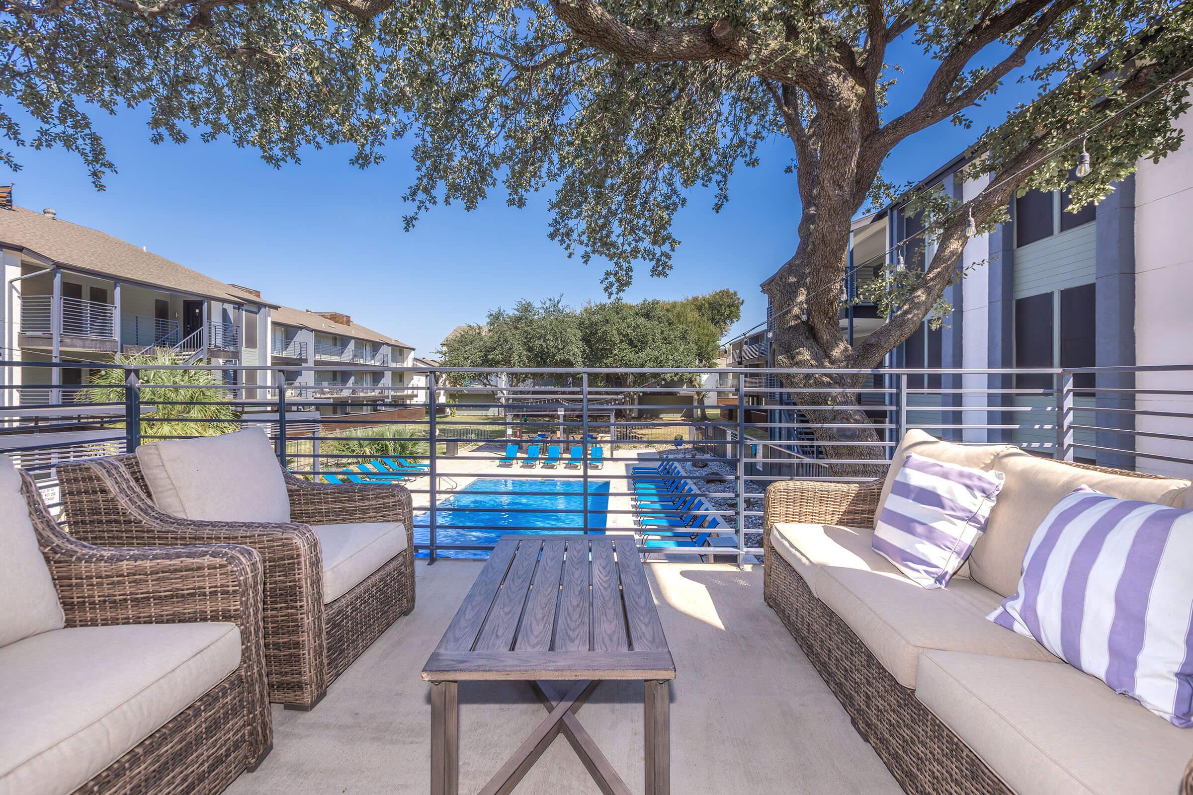 A cozy outdoor seating area featuring two beige wicker couches with striped pillows, positioned on a patio overlooking a swimming pool. Surrounding trees provide shade, and lounge chairs are arranged near the pool. The setting suggests a relaxing and inviting atmosphere for leisure.