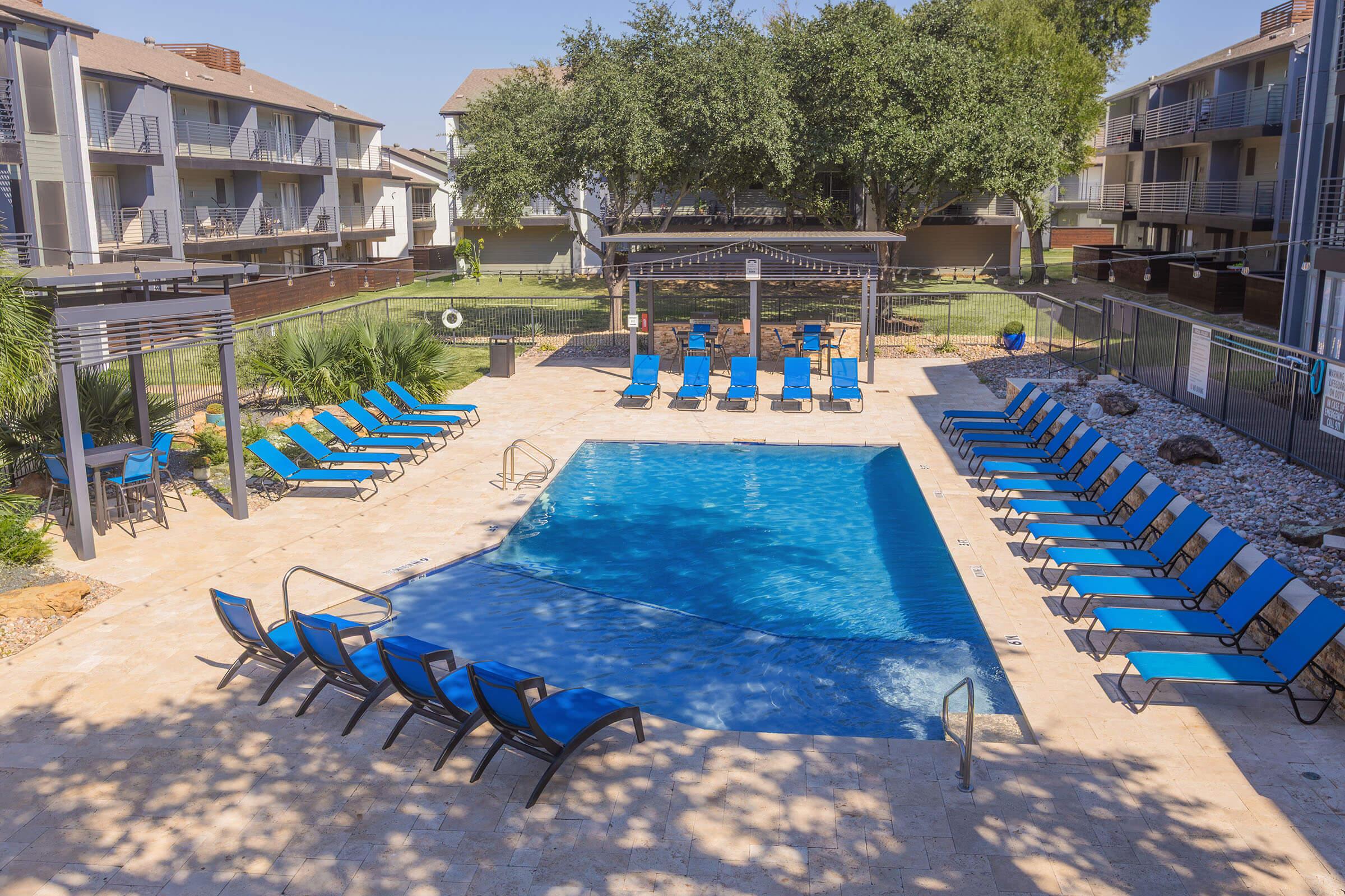 A spacious outdoor pool area featuring a rectangular swimming pool surrounded by blue lounge chairs. Lush green trees provide shade, and there are several multi-story residential buildings in the background. The pool deck is made of stone, creating a relaxing atmosphere for sunbathing and leisure.