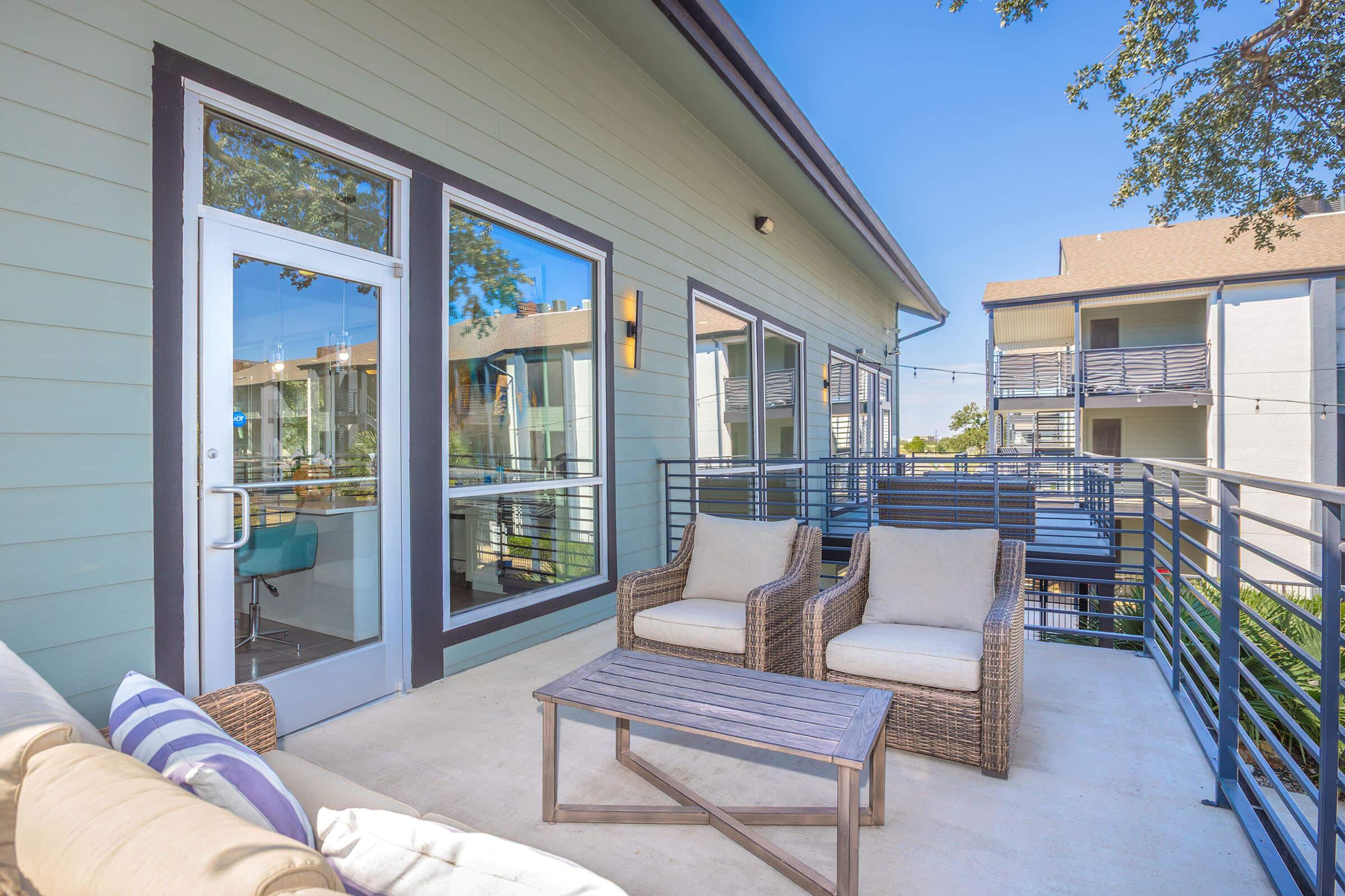 A cozy outdoor patio featuring two wicker chairs and a wooden coffee table, surrounded by greenery. The patio is attached to a light green building with large windows, showcasing a modern architectural design. Bright blue sky and nearby buildings are visible in the background.