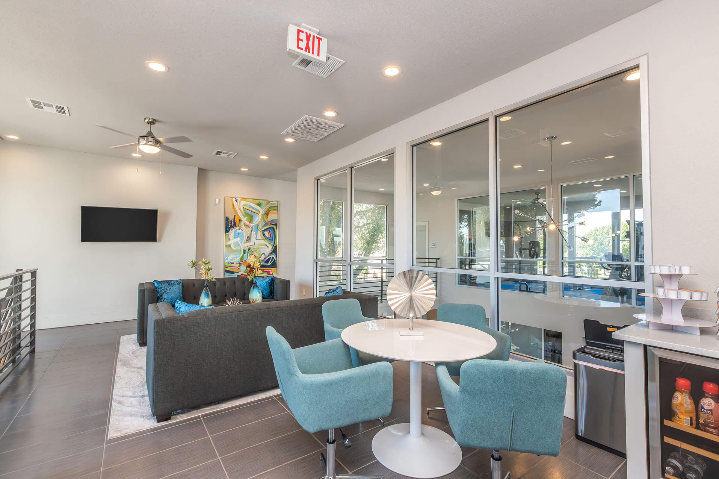 A modern living space featuring a dining table with four teal chairs, a plush couch, and a wall-mounted TV. Large windows provide natural light and a view of the outdoors. Decorative artwork and a stylized centerpiece adorn the table, while a small kitchenette with a beverage station is visible nearby.
