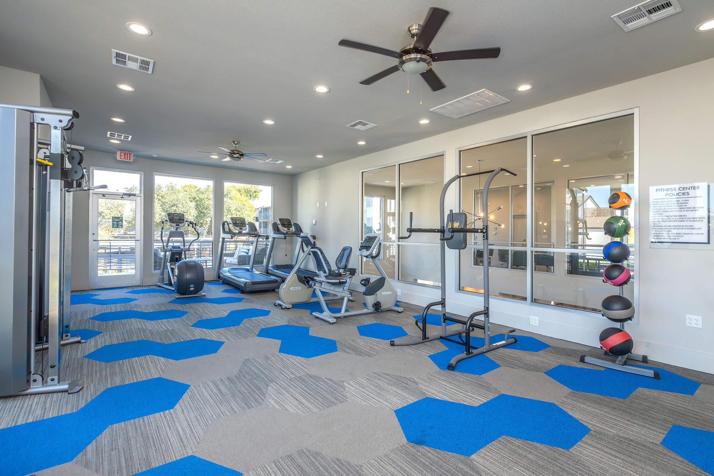 A modern fitness room featuring various exercise equipment, including treadmills, stationary bikes, and strength training machines. The space has blue carpet tiles in hexagonal patterns and large windows allowing natural light. There are also exercise balls on a rack against the wall.