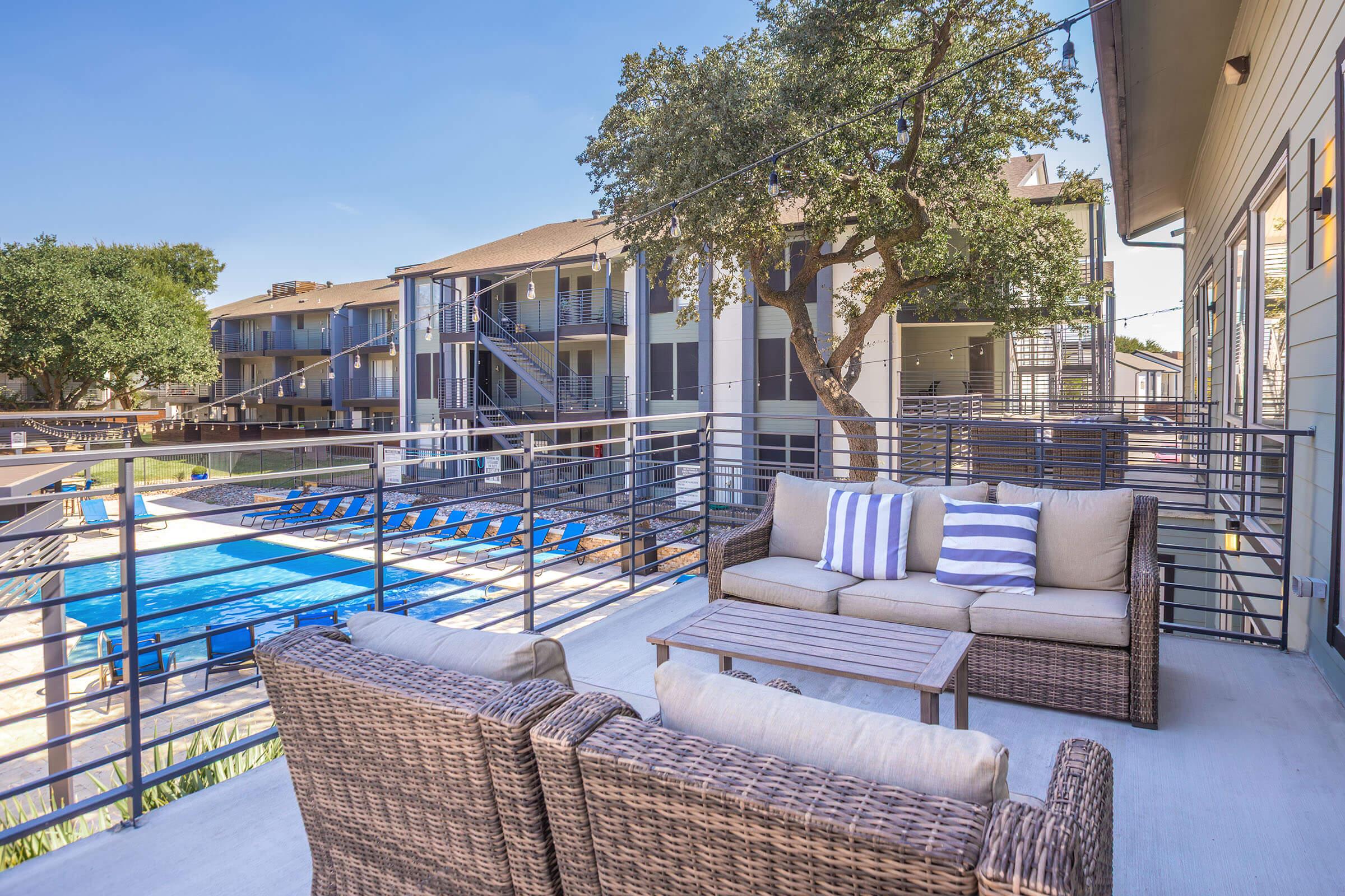 A cozy outdoor lounge area with wicker furniture and decorative pillows overlooks a swimming pool and adjacent buildings. The setting features greenery, blue skies, and a relaxed atmosphere, perfect for enjoying warm weather.