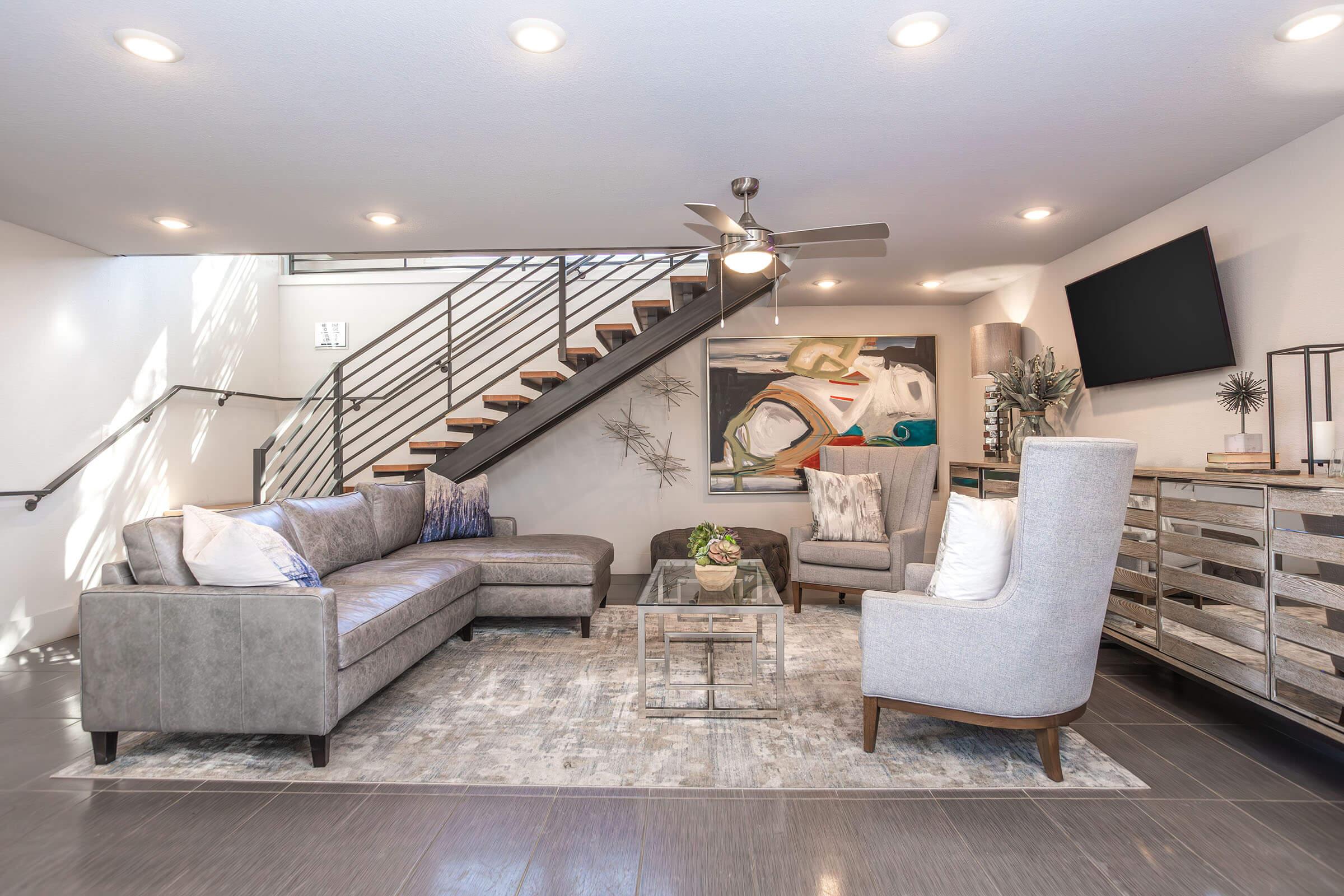 A stylish modern living room featuring a gray sectional sofa, a light gray armchair, and a glass coffee table on a patterned rug. A large abstract painting adorns the wall, with a flat-screen TV mounted nearby. The space also includes a staircase, decorative plants, and contemporary lighting.