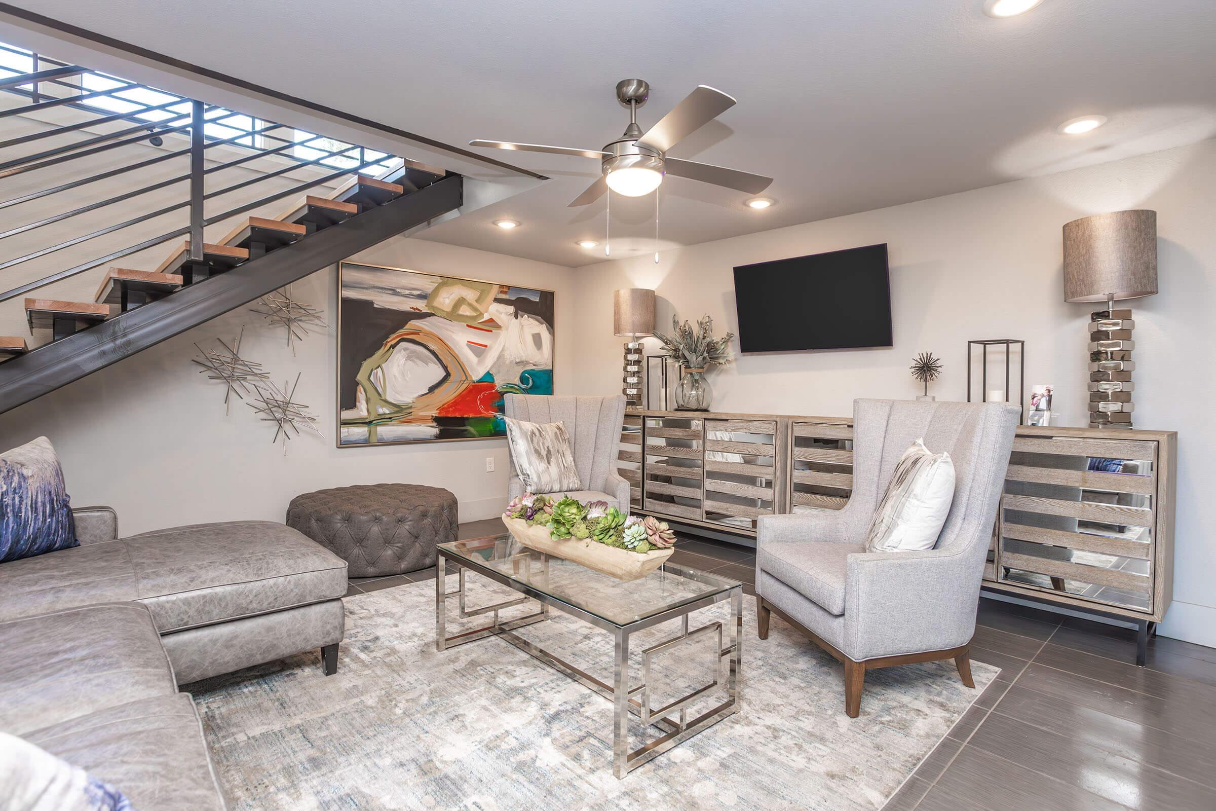 A modern living room featuring a gray sectional sofa, decorative armchairs, and a glass coffee table with a floral centerpiece. The space includes a ceiling fan, abstract wall art, a flat-screen TV mounted on the wall, and stylish lighting fixtures. A staircase is visible in the background.