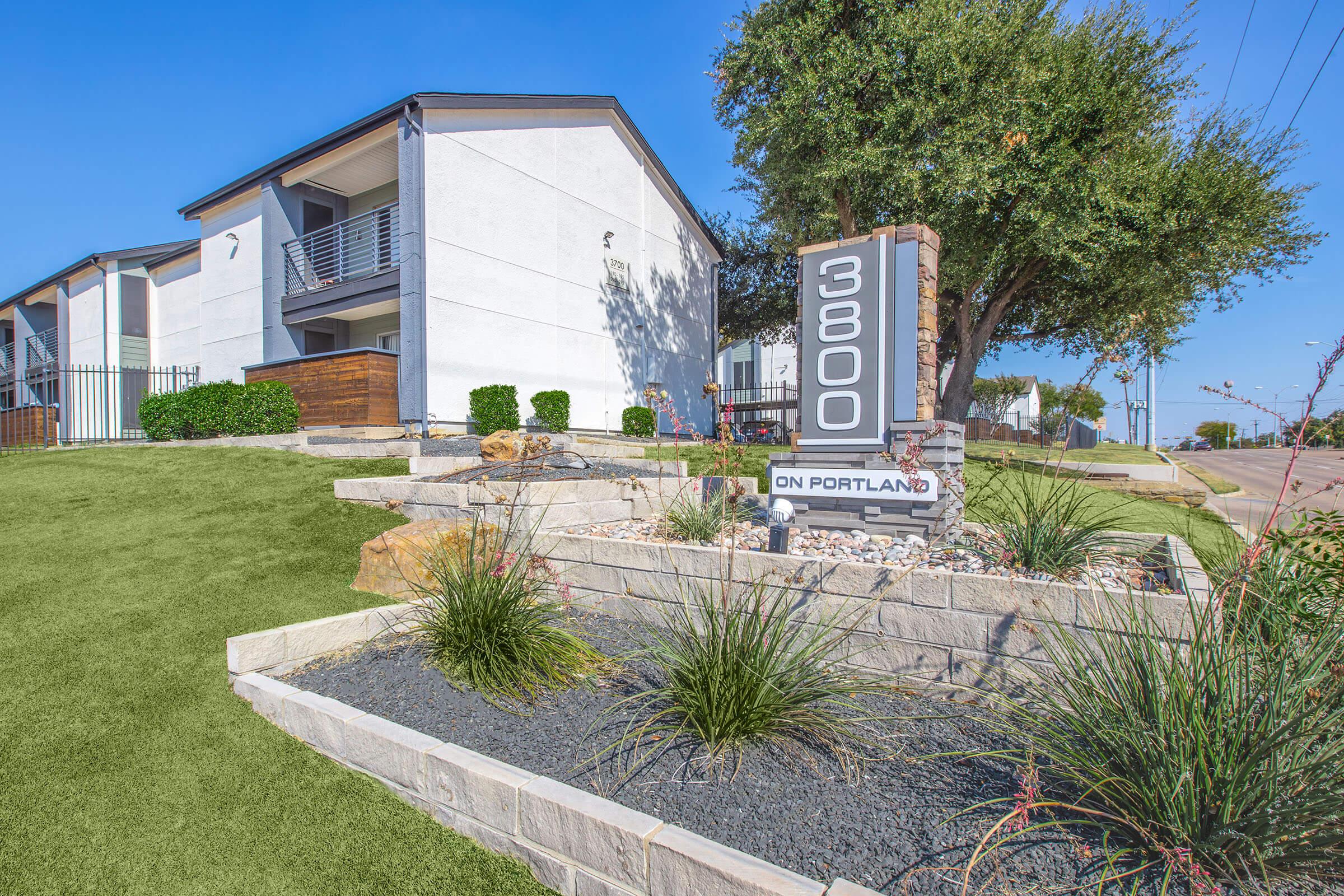 Modern apartment building with a white exterior and multiple balconies. In the foreground, landscaped areas feature ornamental plants and stones, with a sign displaying "3800 ON PORTLAND." A clear blue sky is visible above, contributing to a bright, inviting atmosphere.