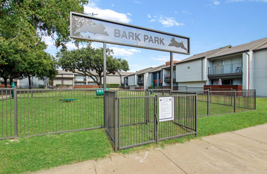 A fenced area labeled "Bark Park" with a sign, surrounded by green grass. Several trees are visible in the background, and nearby buildings can be seen. A notice is posted on the fence, and there is a small play area for dogs inside the park. The sky is clear with a few clouds.
