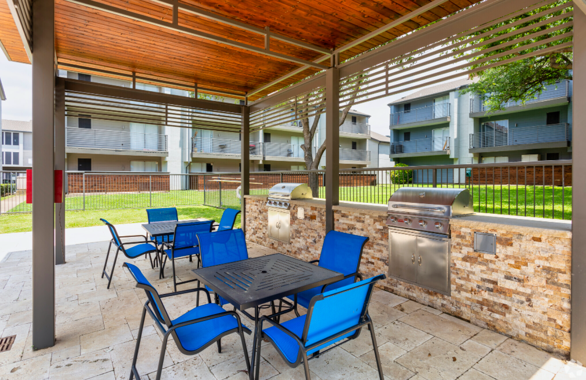 Outdoor shaded patio area featuring a black metal table surrounded by blue chairs. Two stainless steel grills are mounted on a stone wall. The background shows grass and modern apartment buildings with balconies. Ideal setting for gatherings and barbecues.