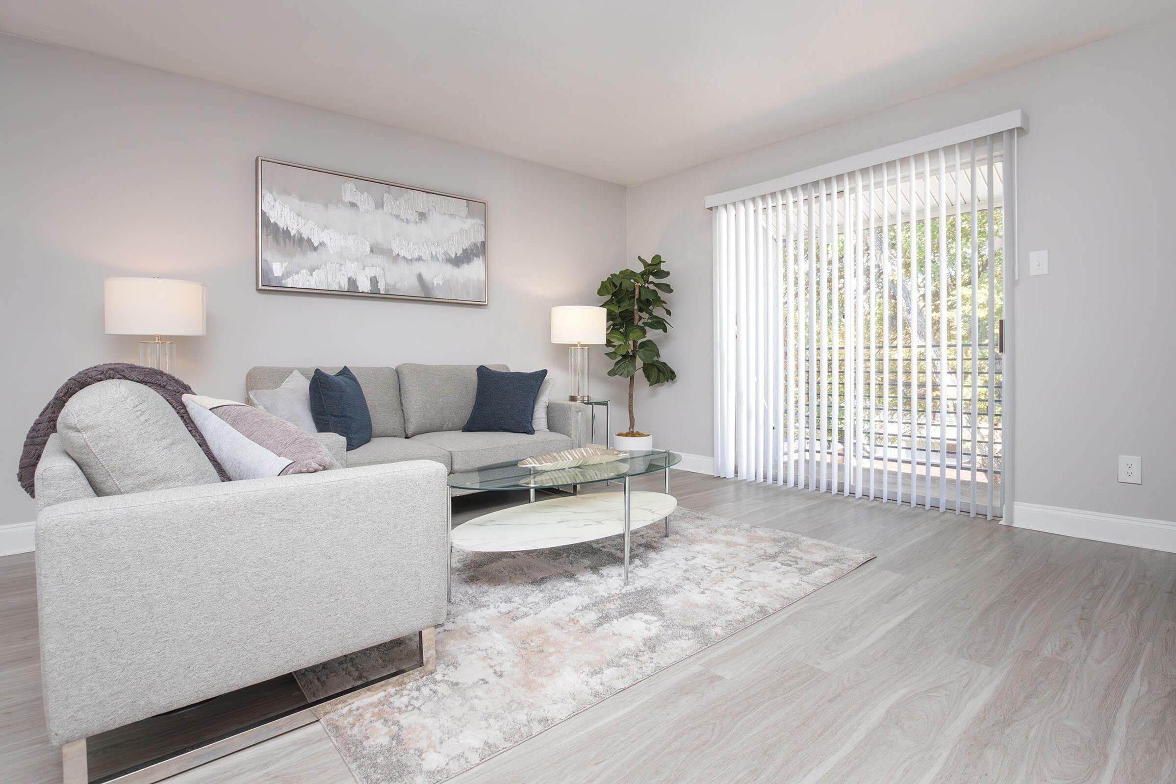 A modern living room featuring a light gray sectional couch adorned with decorative pillows, a glass coffee table, and a soft area rug. There are two lamps providing ambient light, a potted plant for decoration, and a sliding glass door with vertical blinds leading to an outdoor area.