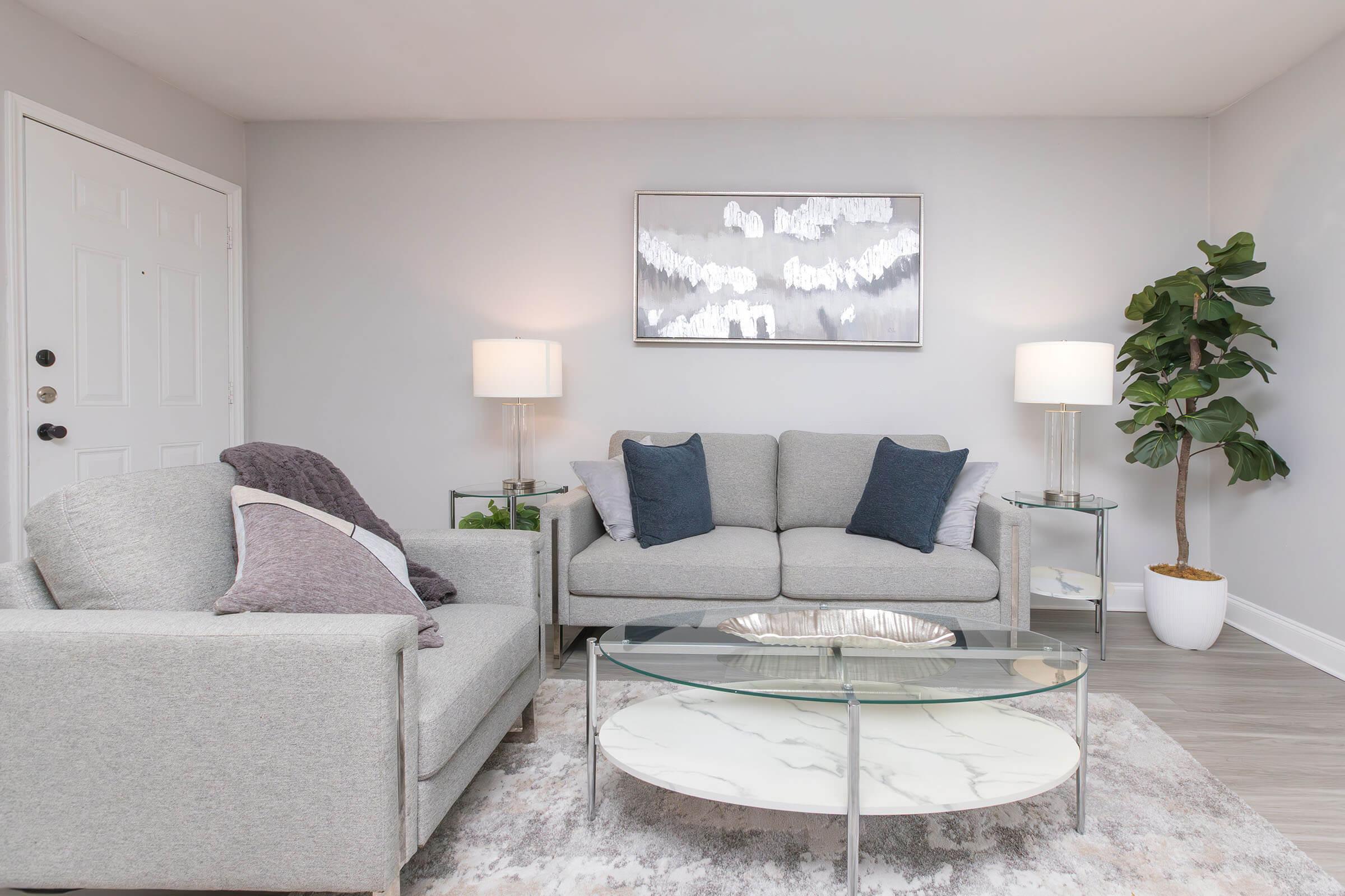 A modern living room featuring a light gray sofa with blue accent pillows, a glass coffee table with a marble top, and a matching armchair. There are two lamps on side tables, a decorative plant in a white pot, and an abstract wall art piece above the sofa, all set against a neutral-colored wall.