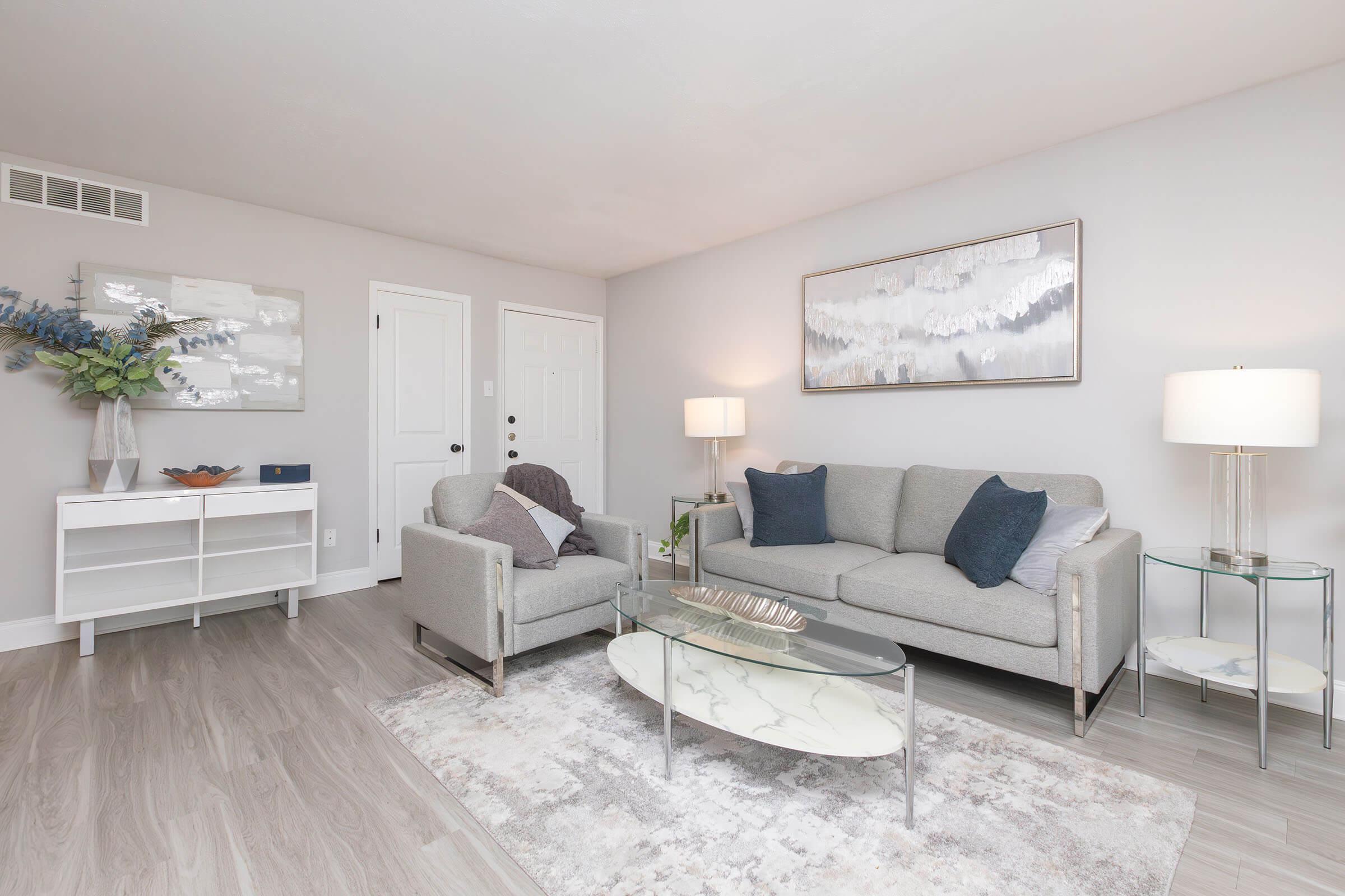 A modern living room featuring a light gray sofa and an armchair, a glass coffee table, and a decorative rug. There are two doors, one leading outside, and artwork hanging on the wall. A small bookshelf and a lamp provide additional decor elements, complemented by a floral arrangement.