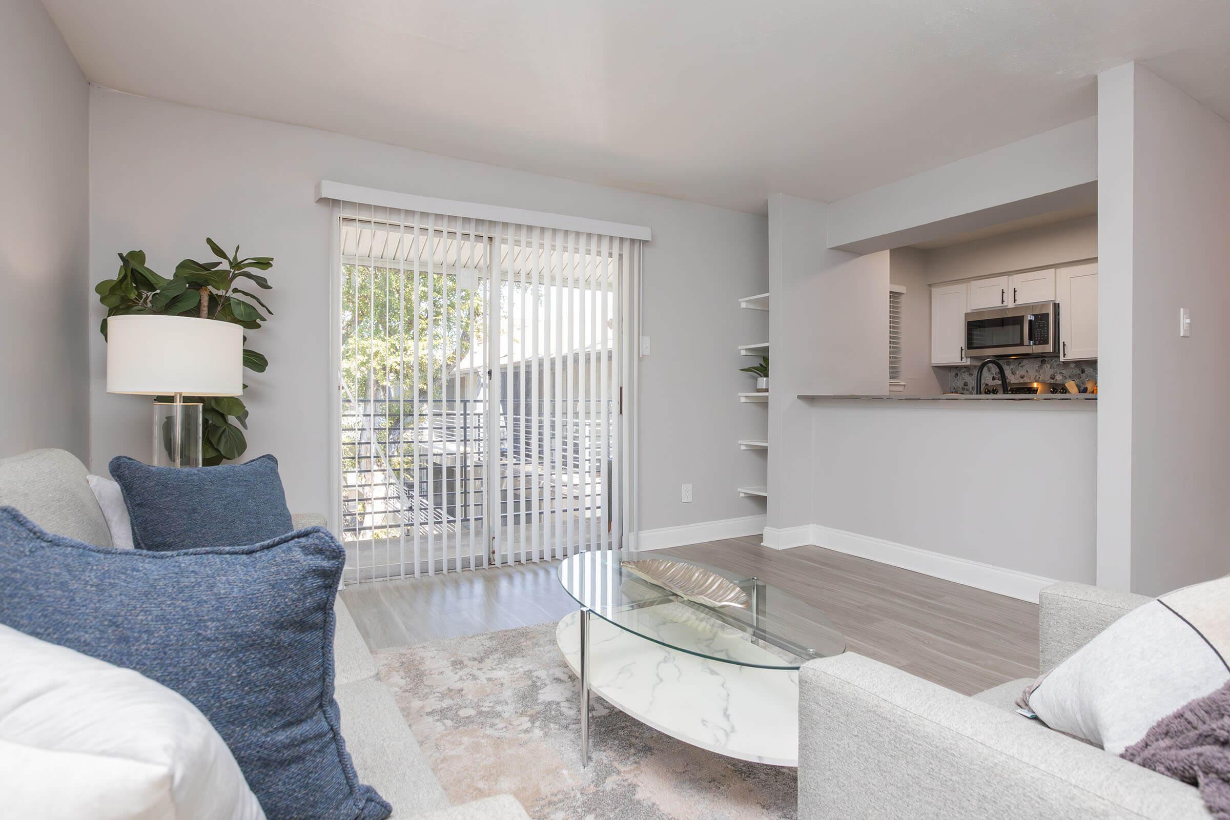 A cozy living room featuring light gray walls, a comfortable sofa with blue cushions, and a glass coffee table. A sliding glass door leads to a balcony, allowing natural light to fill the space. There's a modern kitchen visible in the background, with sleek appliances and open shelving, complemented by a potted plant.