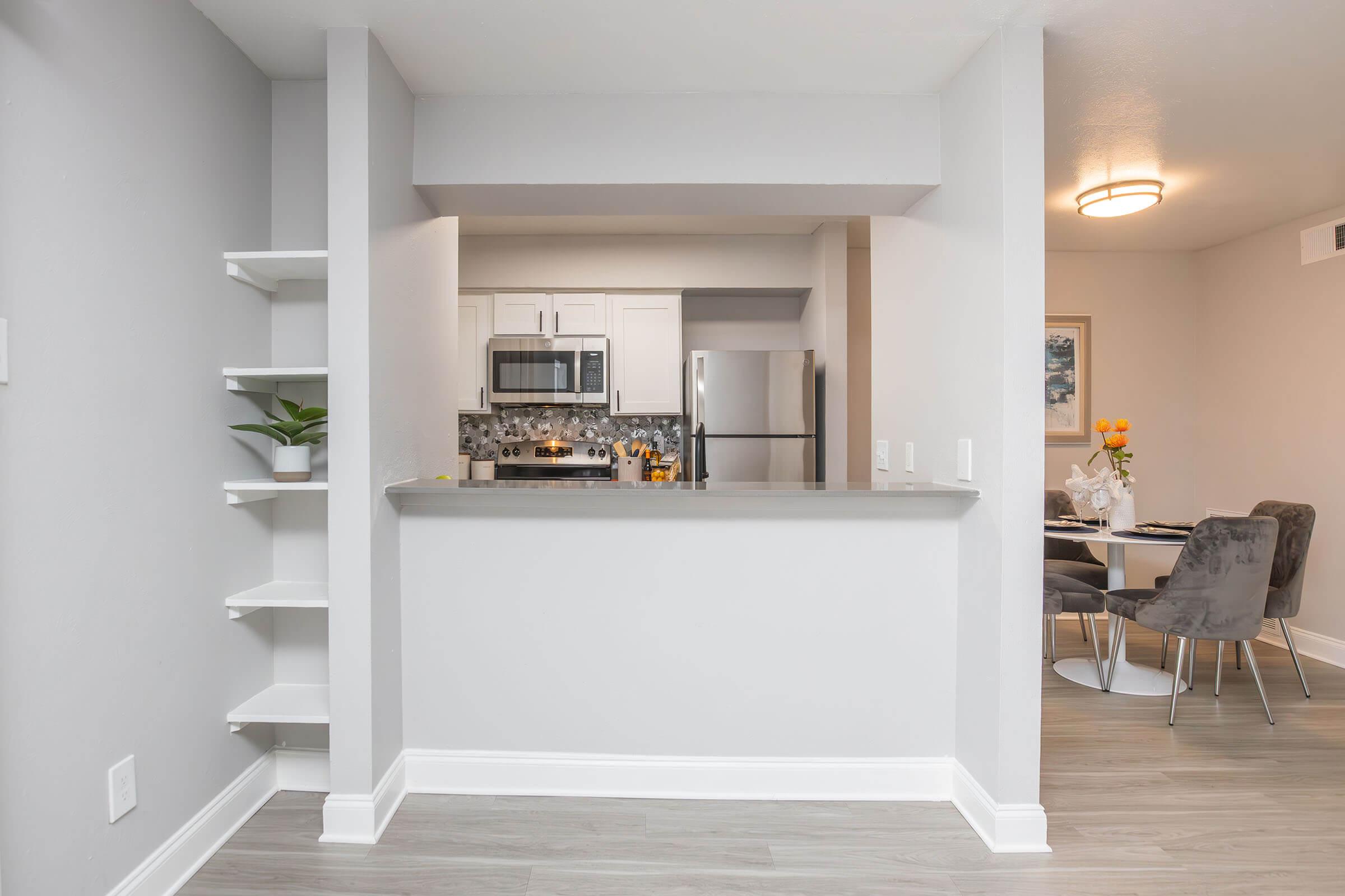 A modern kitchen with gray walls features a small bar area with a plant on a shelf. The kitchen includes white cabinets, stainless steel appliances, and a tile backsplash. In the background, there's a dining area with a round table surrounded by upholstered chairs and a vase of flowers.