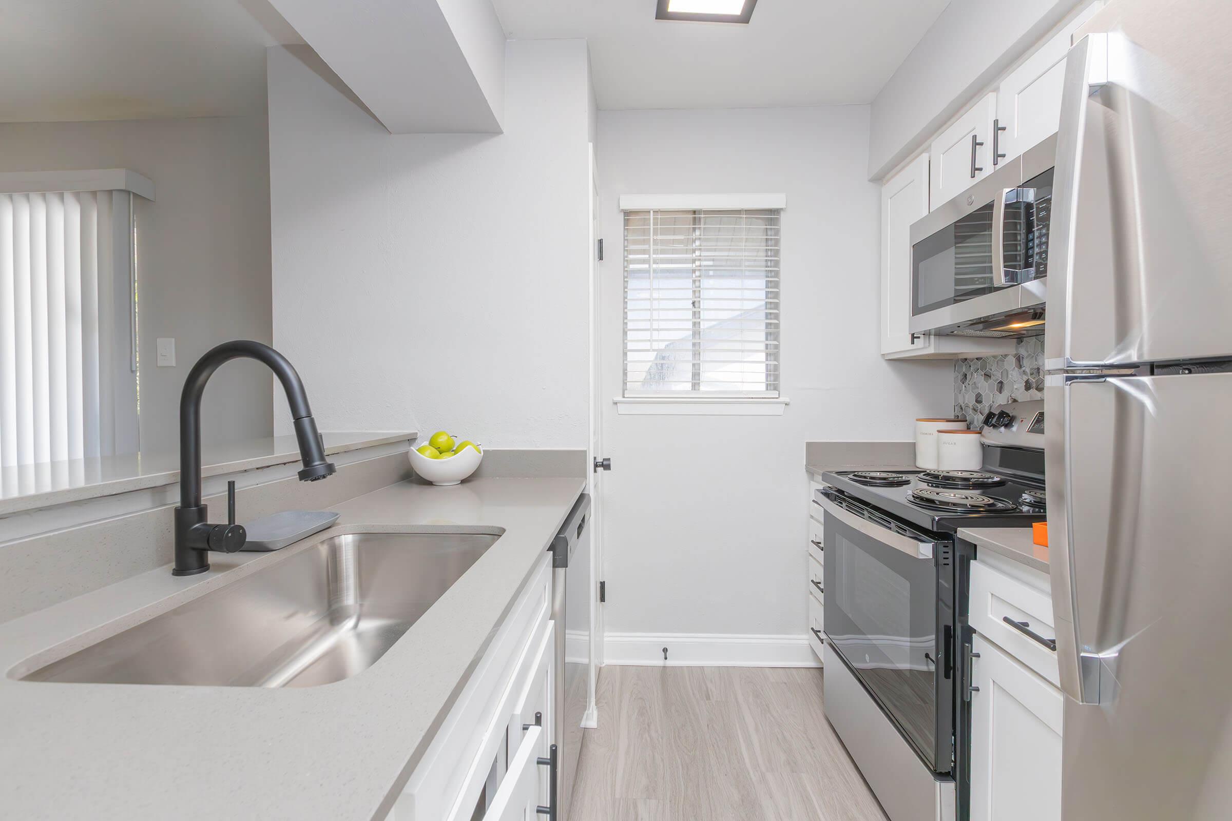 Modern kitchen featuring a stainless steel sink, black faucet, white cabinetry, and appliances including a microwave and oven. There is a bowl of green apples on the countertop, and a window with blinds allows natural light to enter the space. The flooring is light-colored and the overall aesthetic is bright and clean.