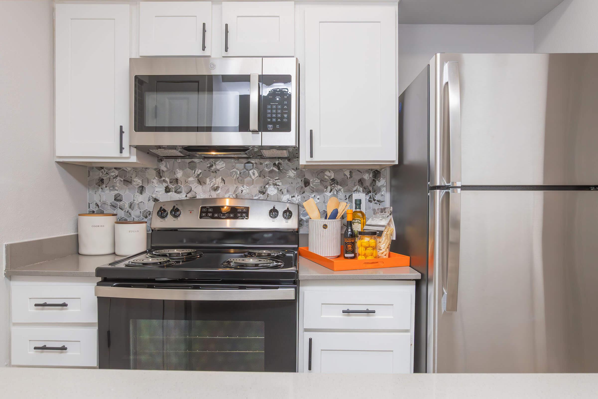 A modern kitchen featuring white cabinets, a silver refrigerator, stainless steel oven, and a microwave. The backsplash is made of decorative tiles. Countertop items include a knife block, spices, and cooking utensils, with an orange tray for organization. Overall, the space is clean and contemporary.