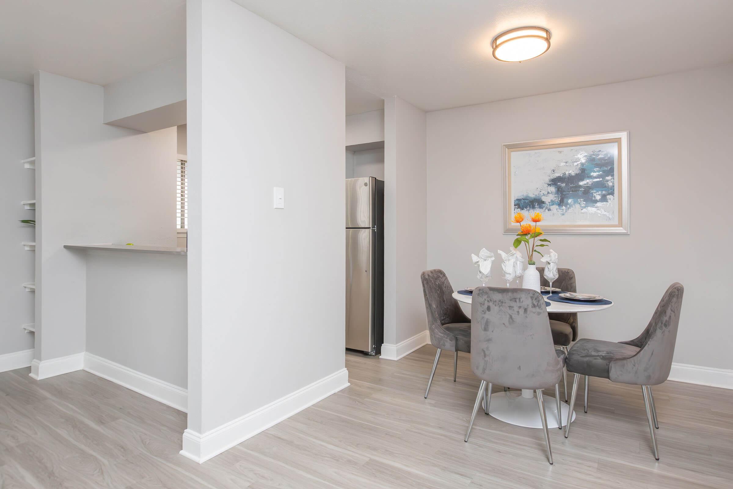 Modern dining area featuring a round table set with a vase of flowers, surrounded by four gray chairs. The walls are painted light gray, and there is a piece of artwork above the table. In the background, a stainless steel refrigerator is visible alongside an open kitchen space with a countertop.