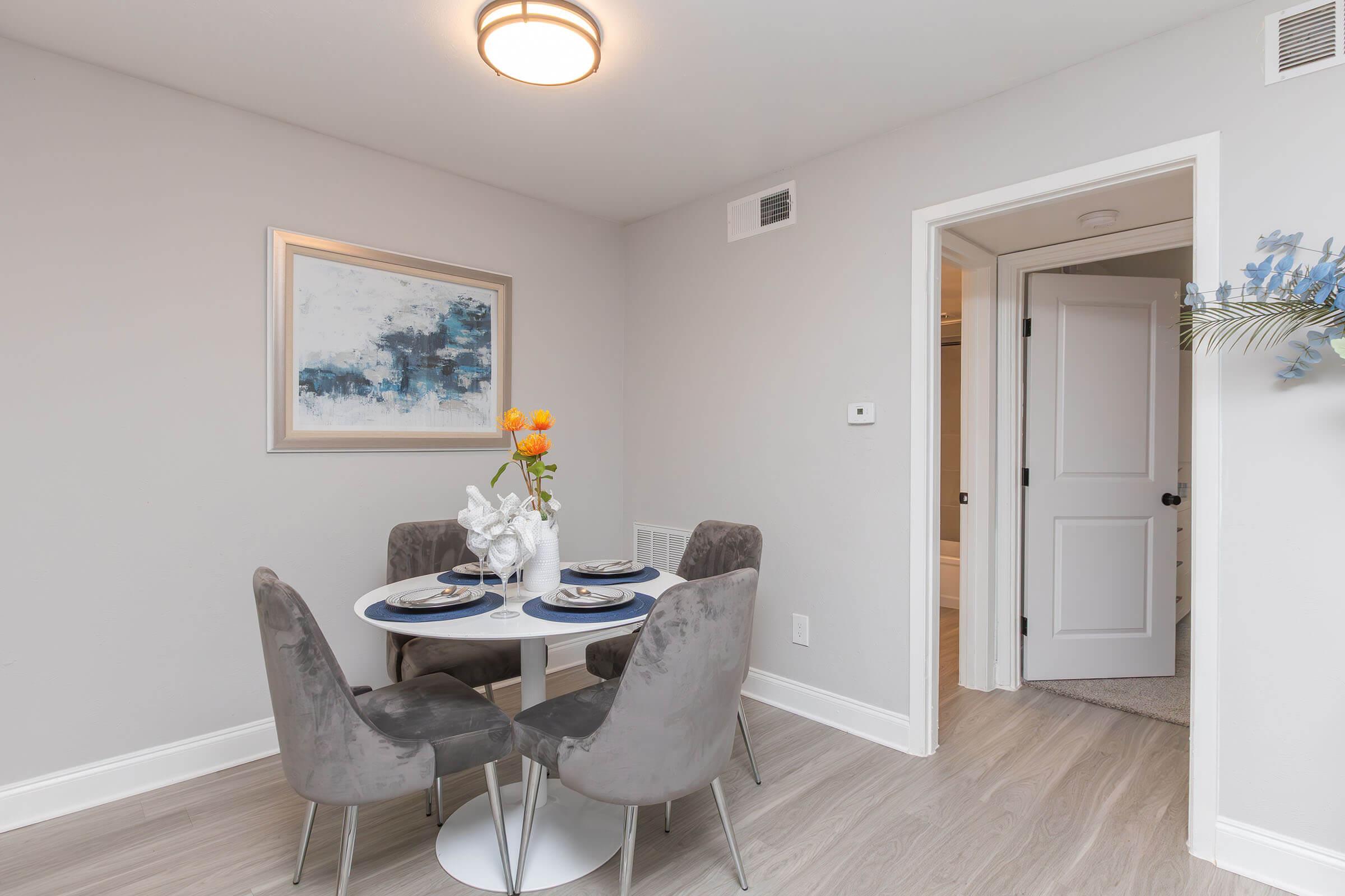 A modern dining area featuring a round table set for two with gray chairs. A floral centerpiece with orange flowers adds a pop of color. The walls are painted light gray and a piece of abstract art hangs on one wall. A door leads to another room in the background, and the flooring is light wood.
